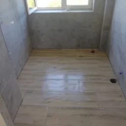 Wykończona łazienka z płytkami imitującymi drewno na podłodze i szarymi płytkami na ścianach. Widoczne okno i otwory na instalacje. Jasne, naturalne światło.