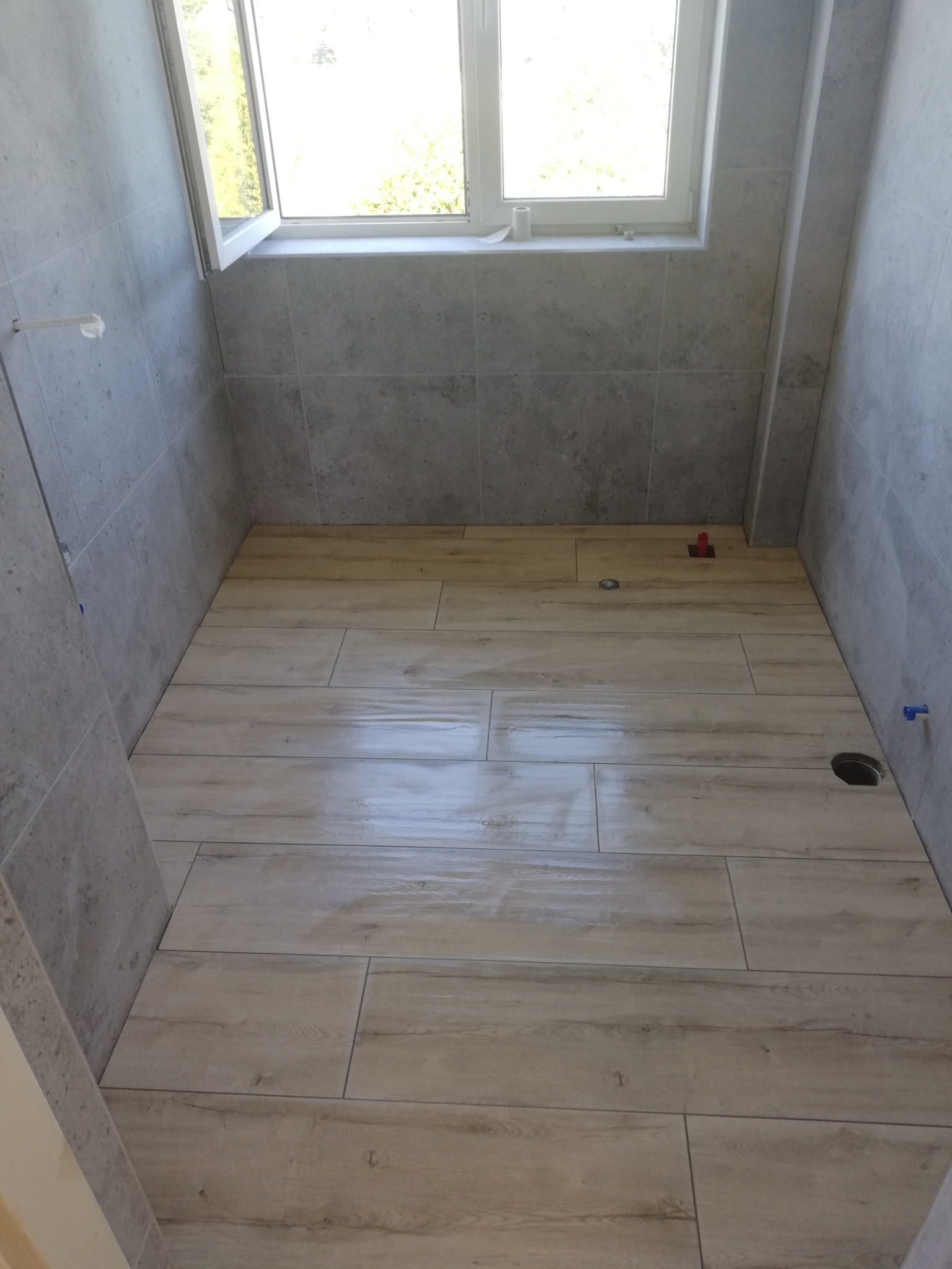 Wykończona łazienka z płytkami imitującymi drewno na podłodze i szarymi płytkami na ścianach. Widoczne okno i otwory na instalacje. Jasne, naturalne światło.