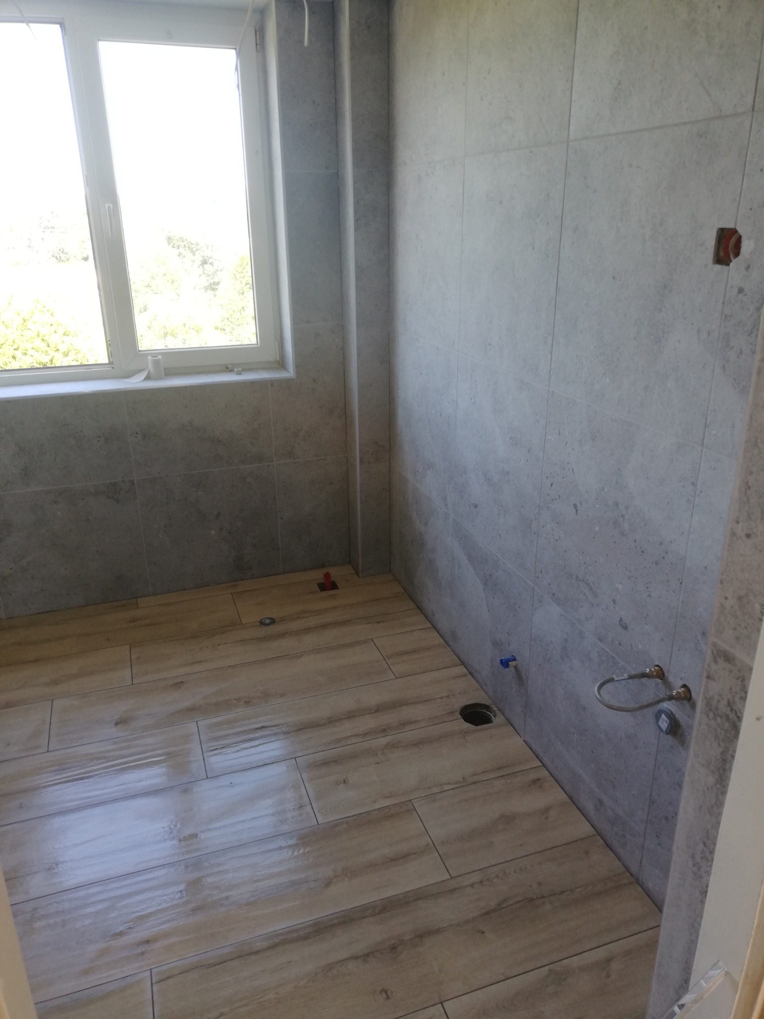 Wykończona łazienka z szarymi płytkami ściennymi i podłogą imitującą drewno. Widoczne przyłącza wodne i otwór kanalizacyjny. Jasne okno doświetla wnętrze.