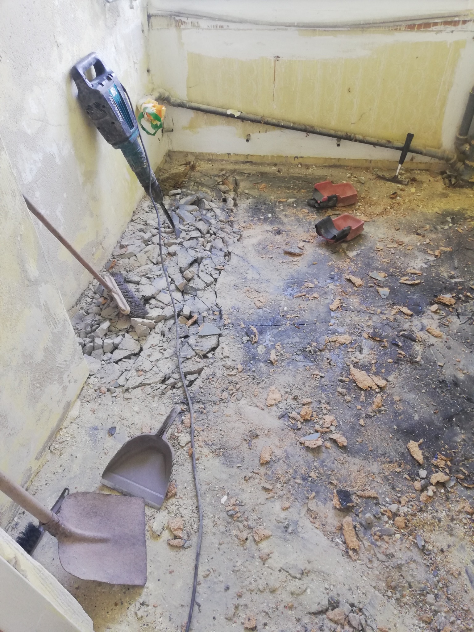 Demontaż podłogi w pomieszczeniu. Na podłodze gruz, narzędzia: młot udarowy, łopata i szufelka. Widoczne rury i fragmenty ścian z resztkami tapet.