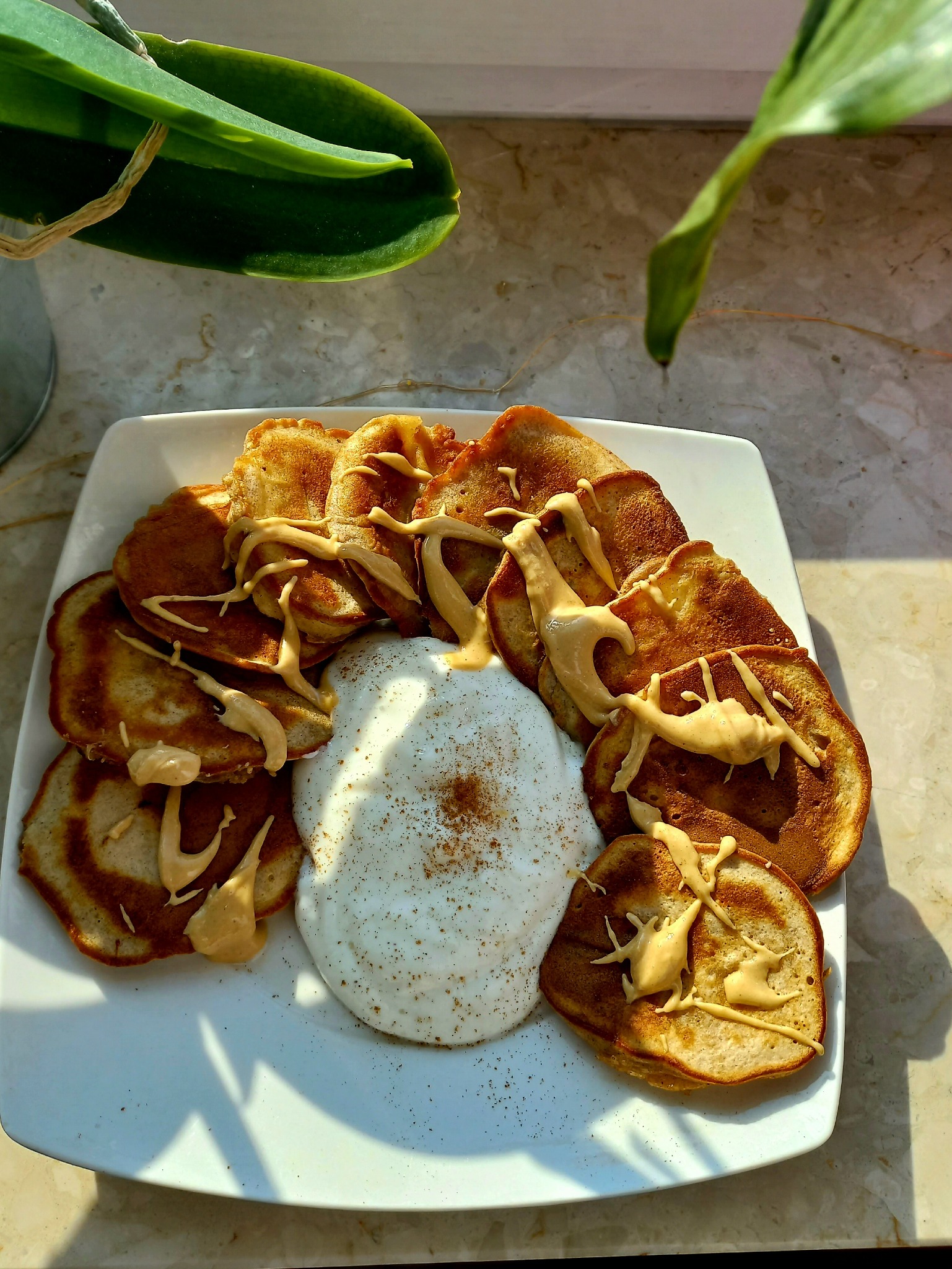Stos bananowych placuszków polanych kremem orzechowym i porcją bitej śmietany posypanej cynamonem, na białym kwadratowym talerzu, z liściem orchidei w tle.