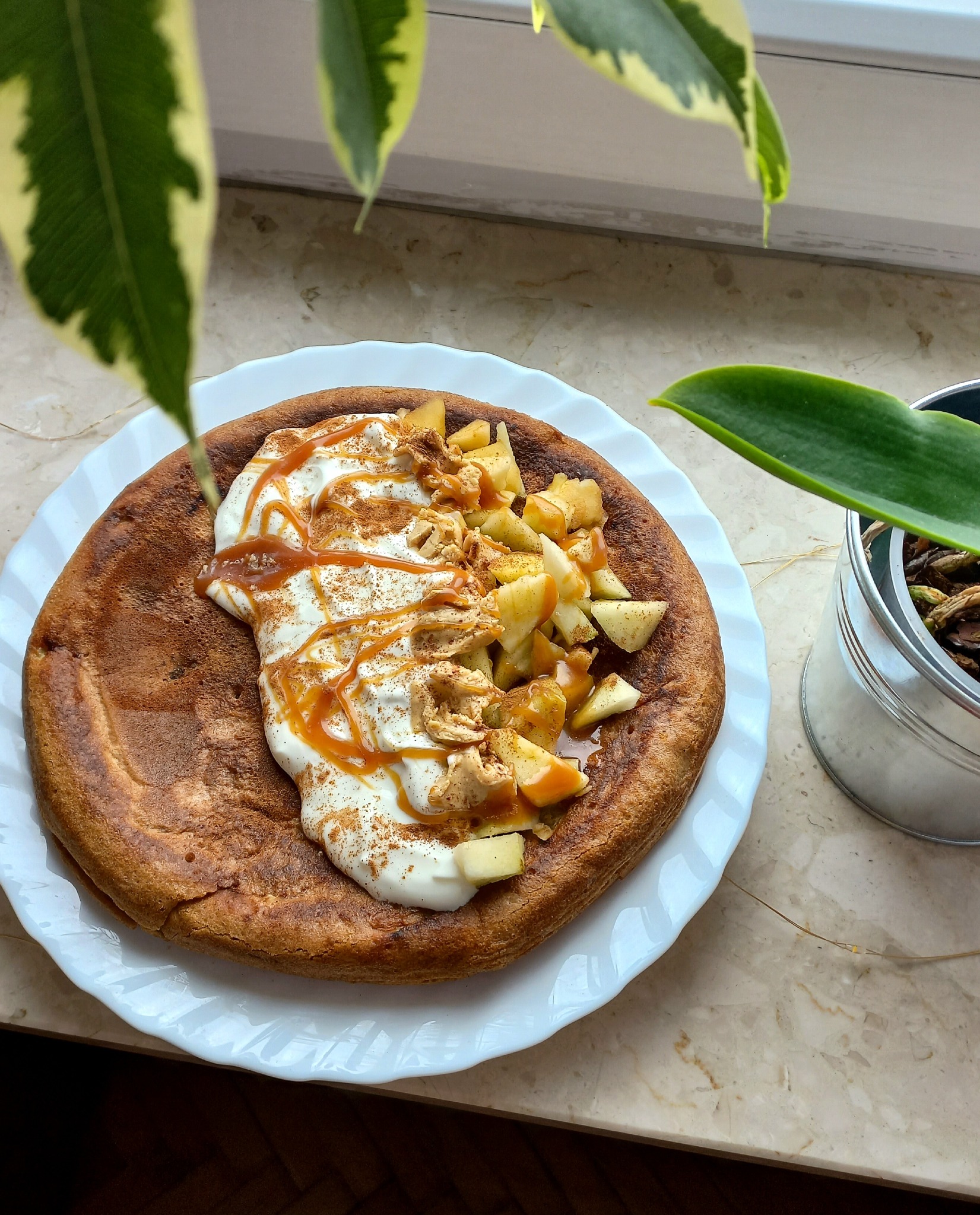 Puszysty naleśnik holenderski na białym talerzu, polany kremem, jabłkami i karmelem, posypany cynamonem, z roślinami w tle.