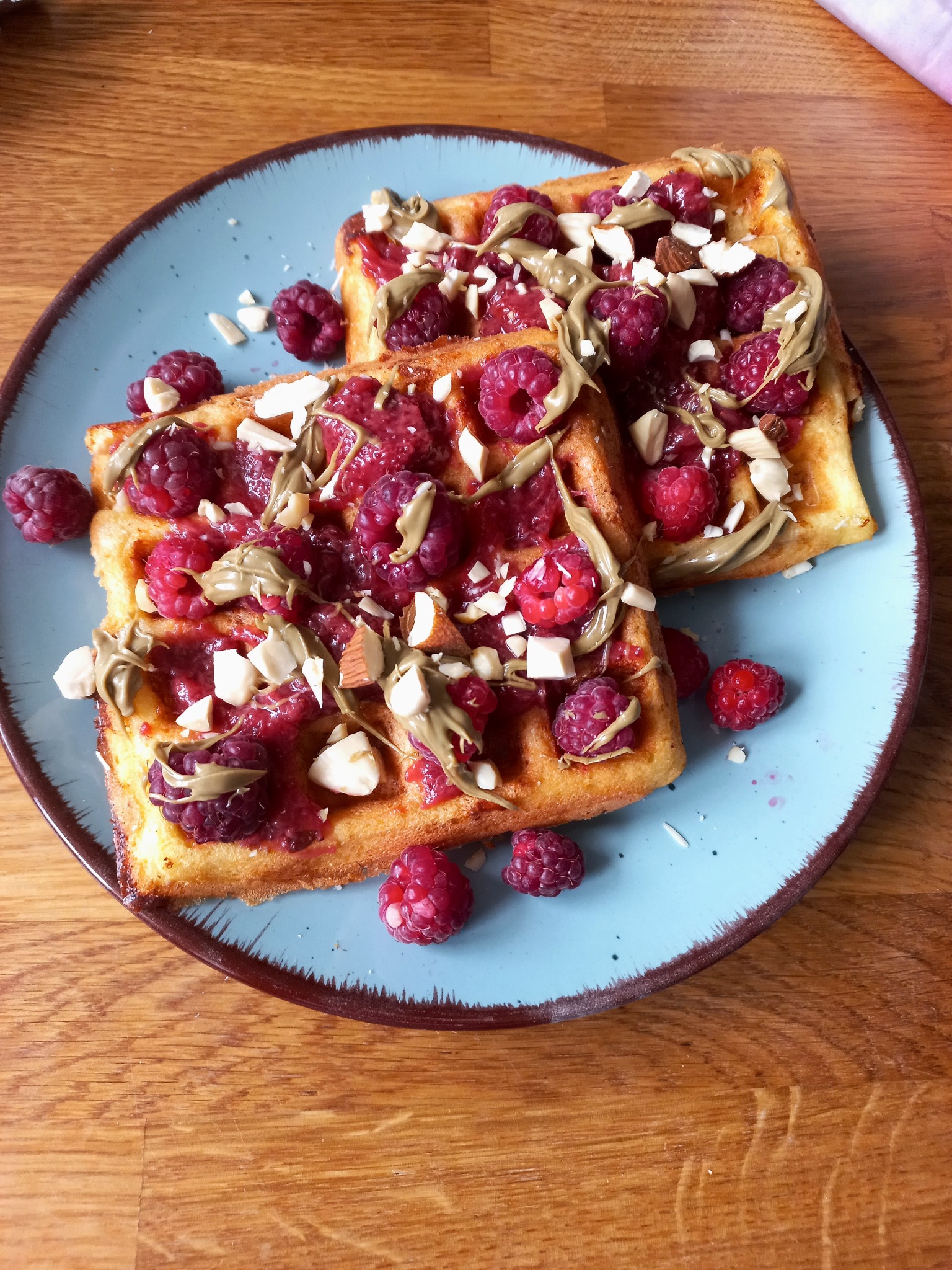 Dwa kwadratowe gofry na błękitnym talerzu z brązową obwódką, polane dżemem malinowym, kremem pistacjowym i posypane płatkami migdałów, kilka malin obok na drewnianym blacie.