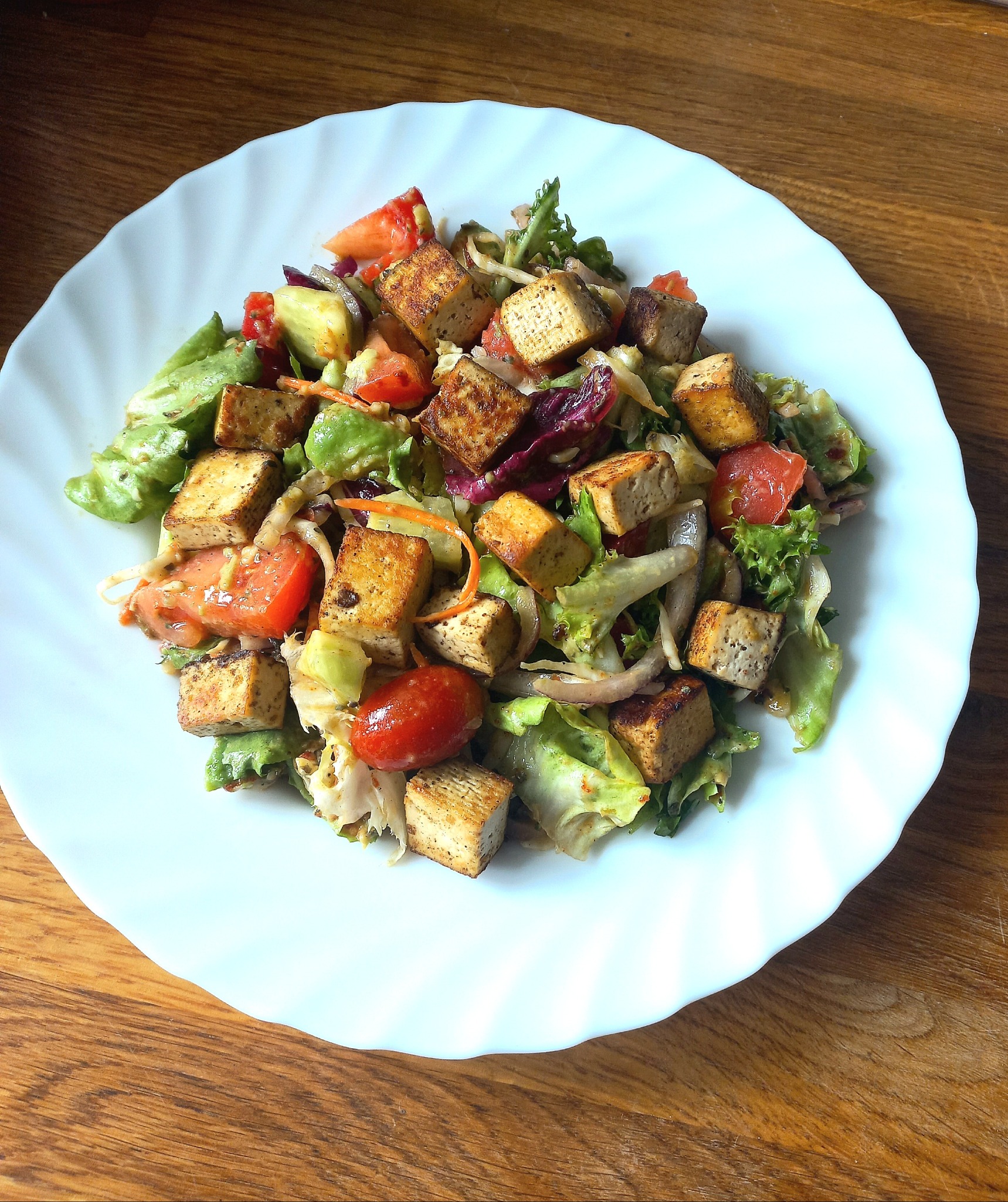 Sałatka z tofu, pomidorów i mieszanki sałat na białym talerzu z falistym brzegiem, ułożonym na drewnianym stole.