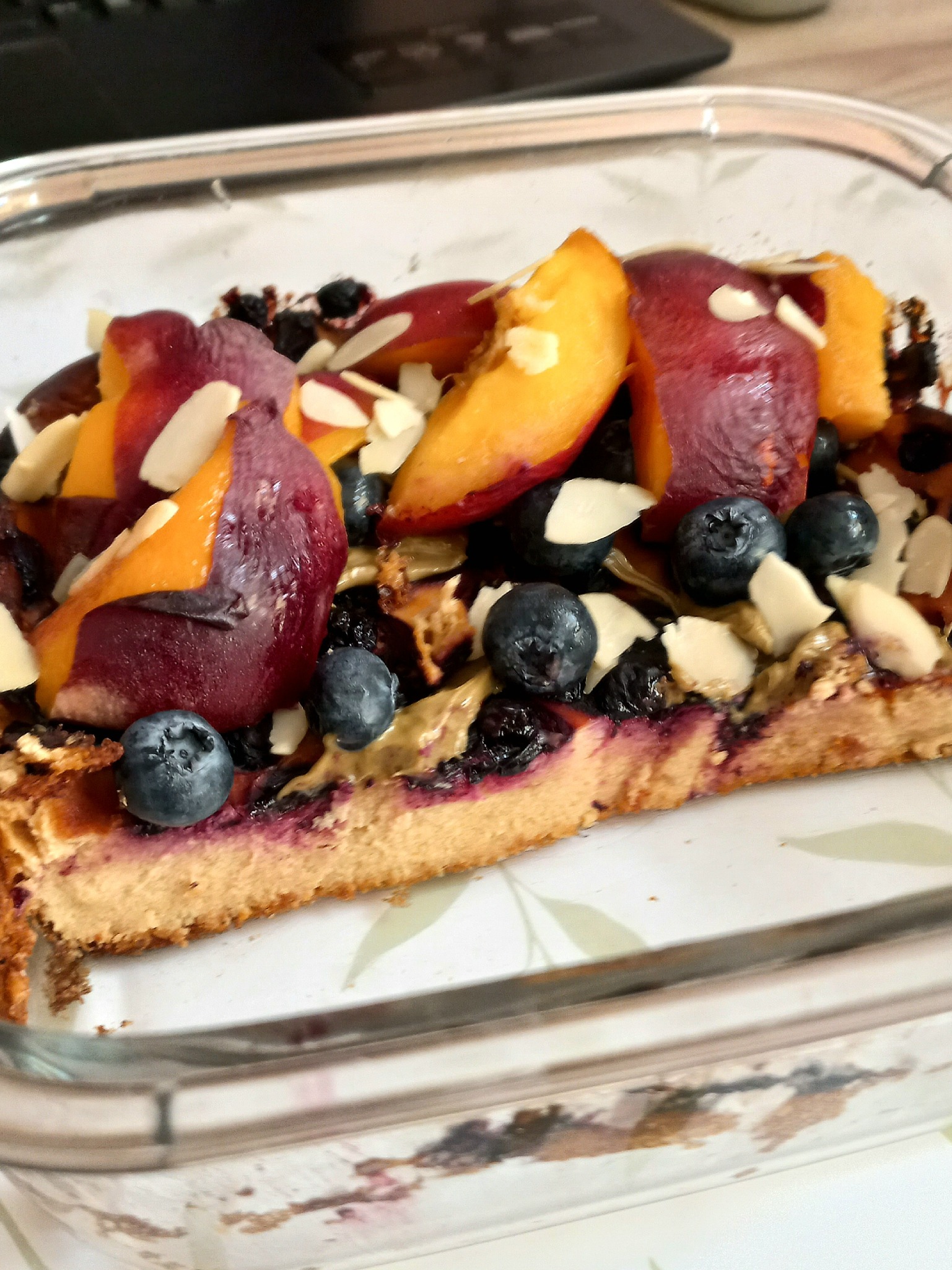 Kawałek ciasta z owocami: śliwki, brzoskwinie, jagody i płatki migdałów, widoczne w szklanym naczyniu do pieczenia.