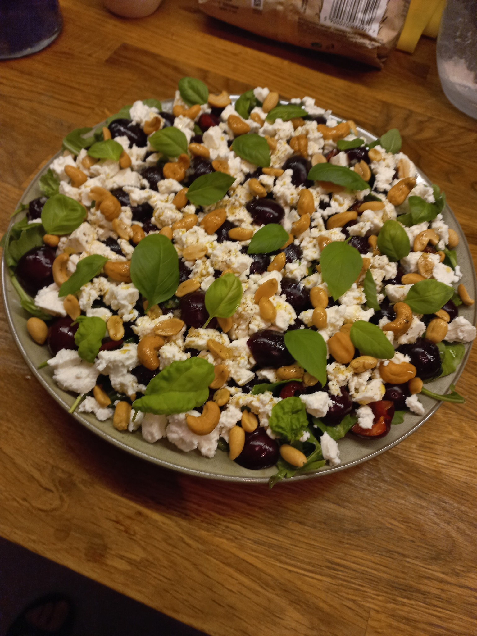 Sałatka z rukoli, sera feta, ciemnych wiśni, orzechów nerkowca i listków bazylii na okrągłym talerzu, ułożona na drewnianym stole.