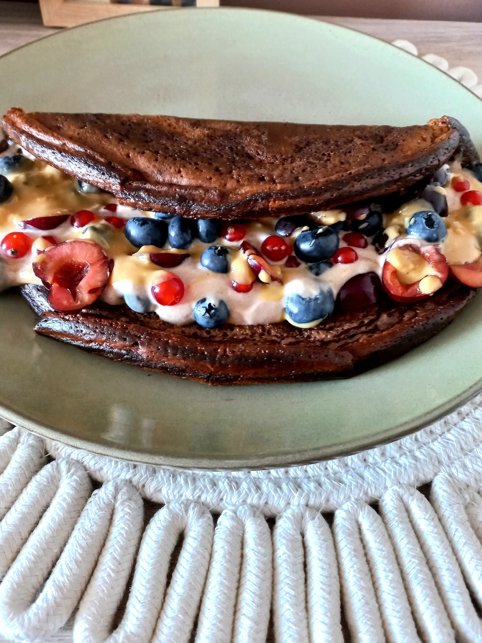 Ciemny naleśnik przełożony kremem, borówkami, wiśniami i czerwoną porzeczką, polany sosem na zielonym talerzu z białym podkładem.