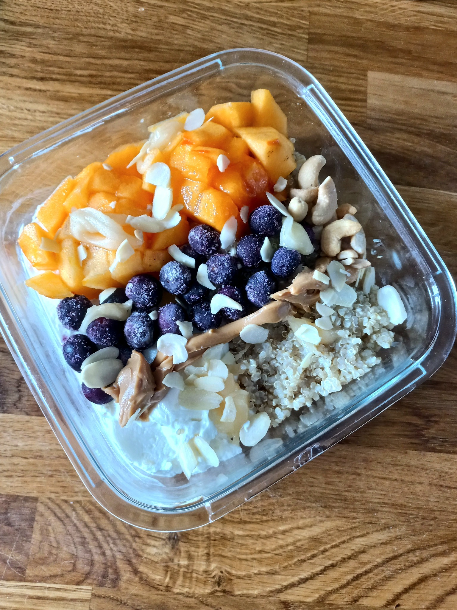 Zdrowe śniadanie w szklanym pojemniku: mango, borówki, orzechy nerkowca, płatki migdałów, quinoa i krem orzechowy na bazie jogurtu.
