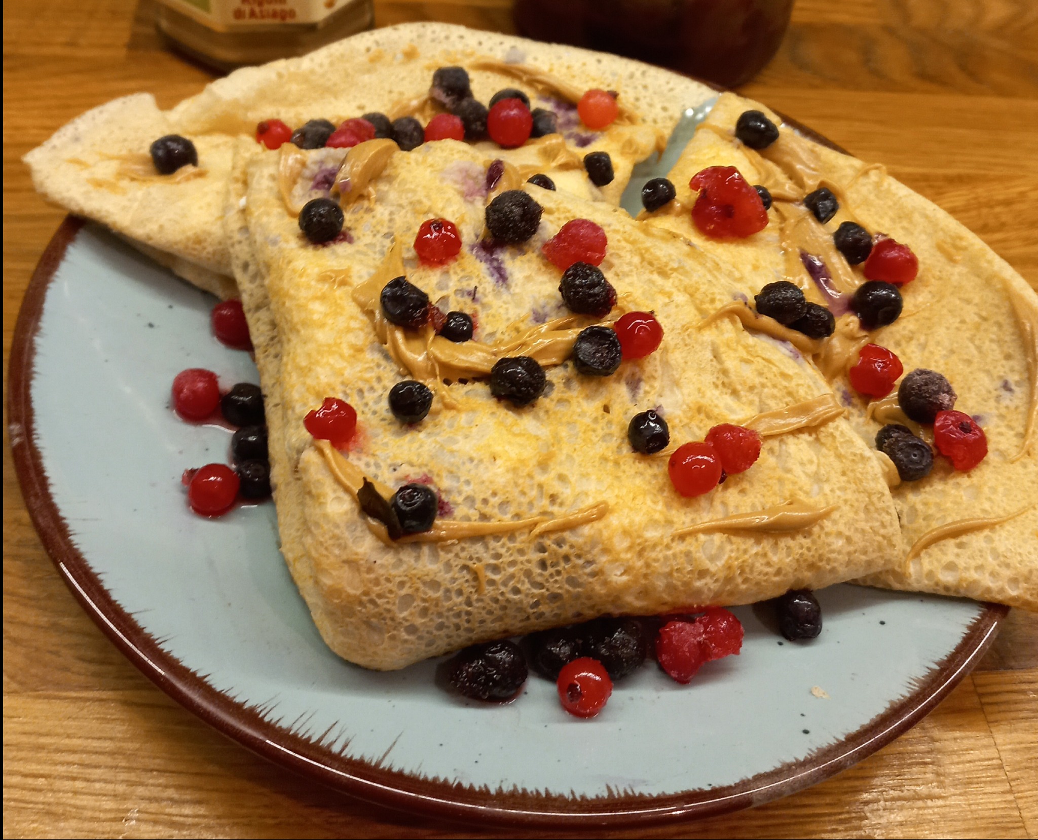Naleśniki z masłem orzechowym, borówkami i czerwoną porzeczką na turkusowym talerzu z brązową obwódką, ułożone na drewnianym stole.
