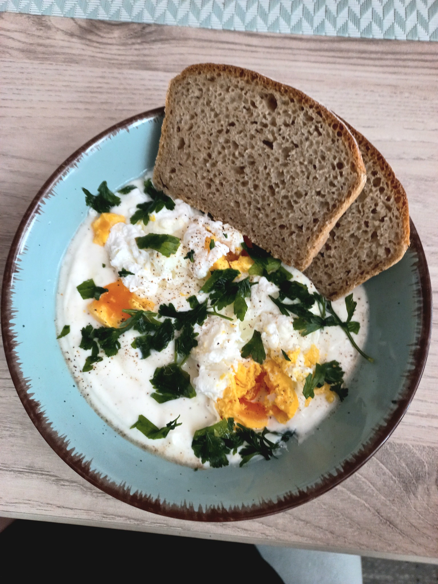 Śniadanie: jajka w koszulce w jogurcie naturalnym z posiekaną natką pietruszki i dwoma kromkami chleba razowego na turkusowym talerzu z brązową obwódką.