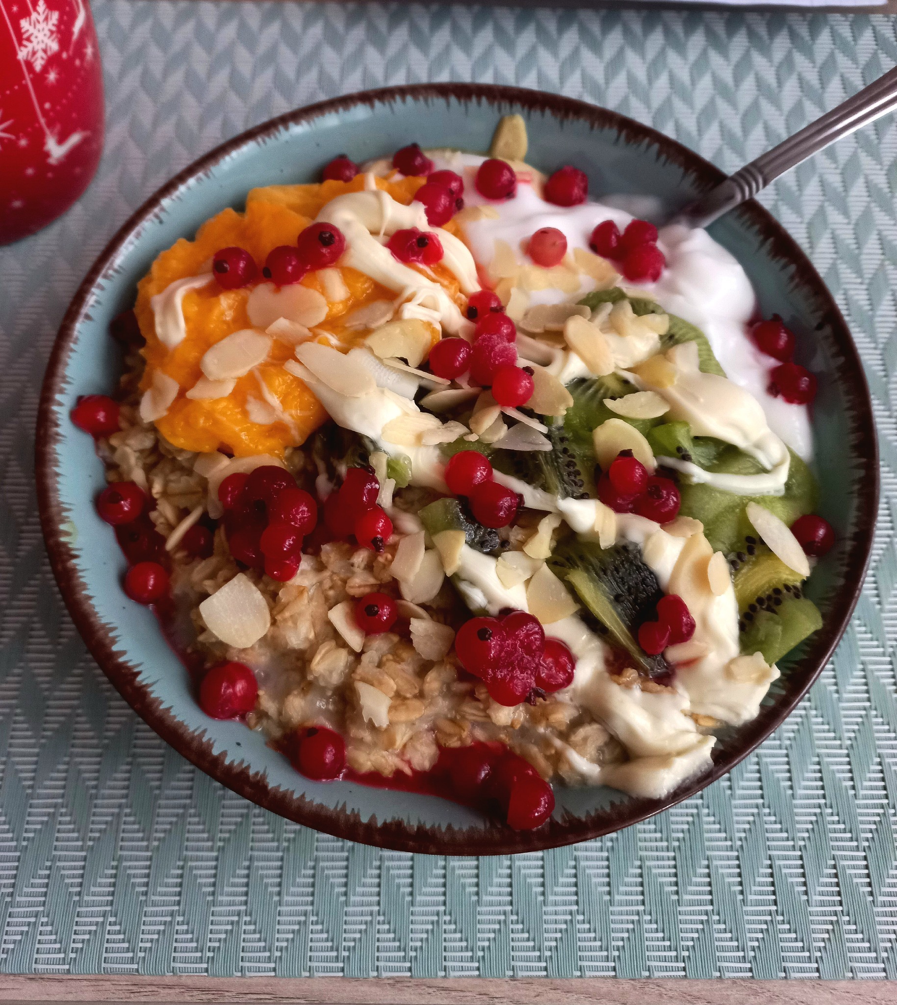 Miska owsianki z pomarańczą, kiwi, jogurtem, płatkami migdałów i czerwoną porzeczką na niebieskiej podkładce.