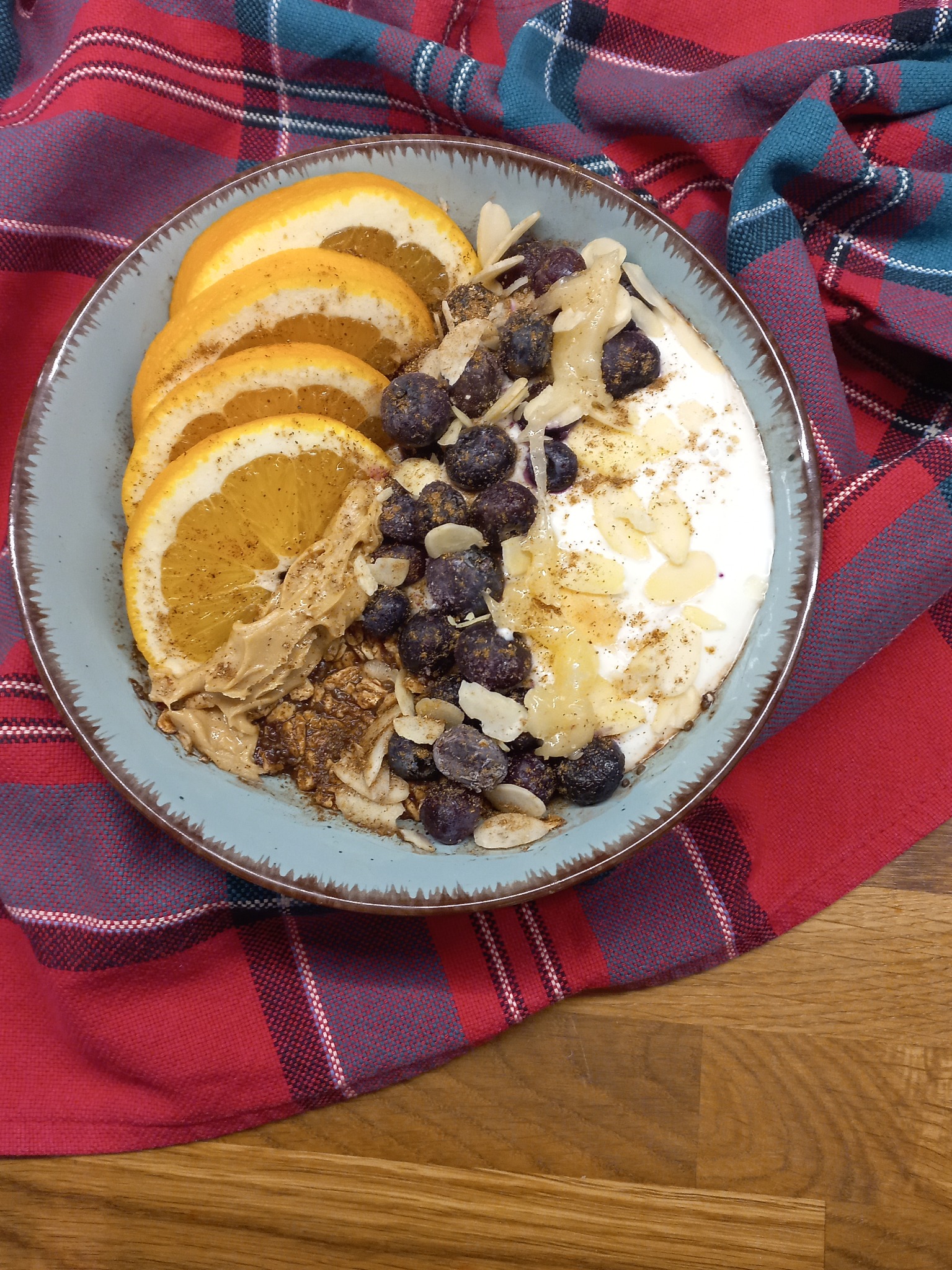 Porcja jogurtu z plasterkami pomarańczy, borówkami, masłem orzechowym i płatkami migdałów w niebieskiej miseczce, na czerwono-niebieskiej kraciastej ściereczce na drewnianym stole.