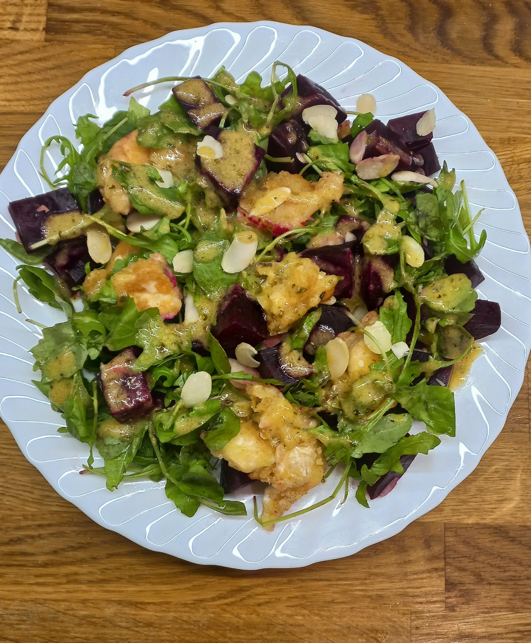 Sałatka z buraków, rukoli, smażonego sera i płatków migdałów, polana sosem musztardowo-miodowym, podana na białym talerzu na drewnianym stole.