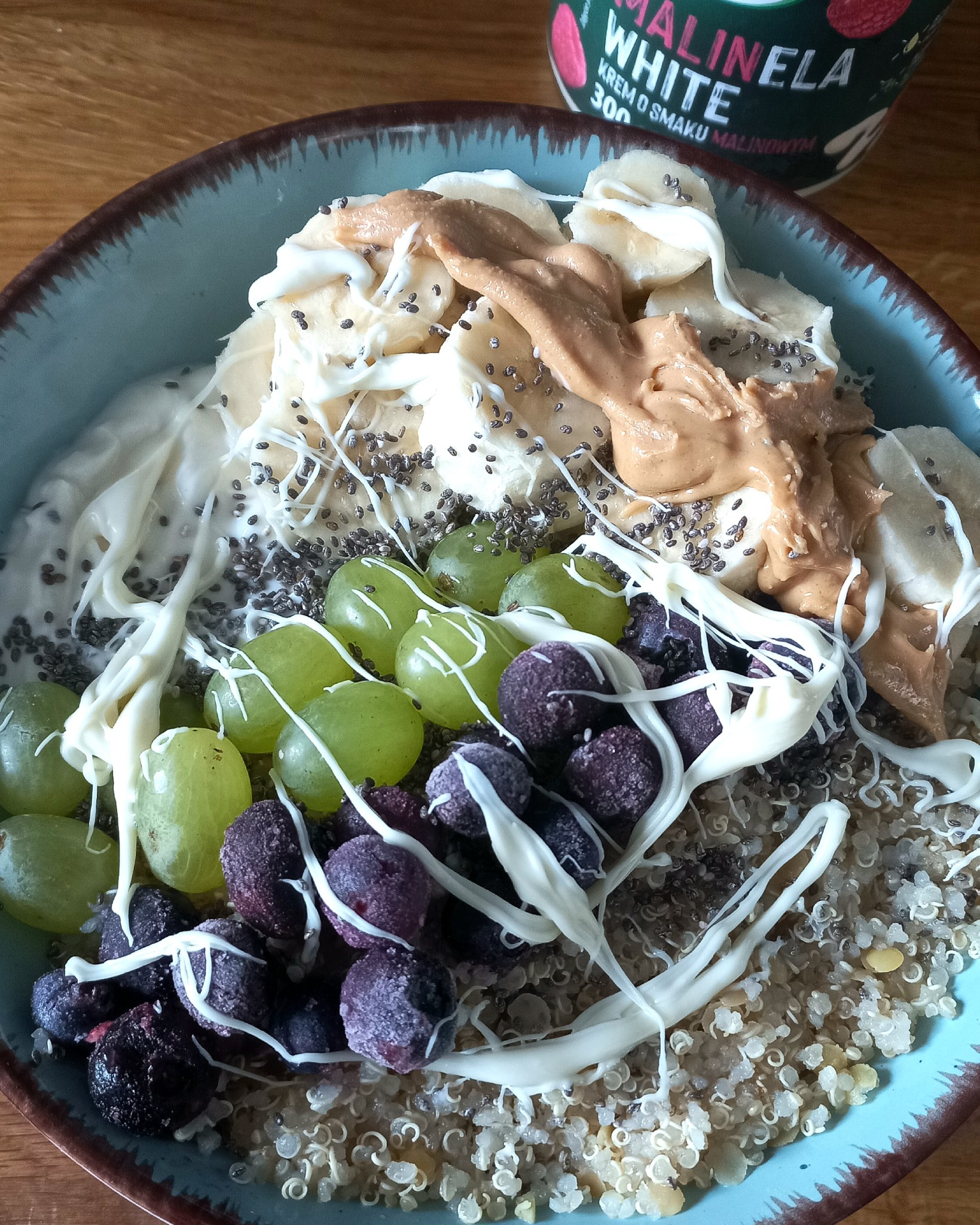 Miska z komosą ryżową, bananem, winogronami, borówkami, masłem orzechowym, nasionami chia i sosem Malinela White na drewnianym stole.