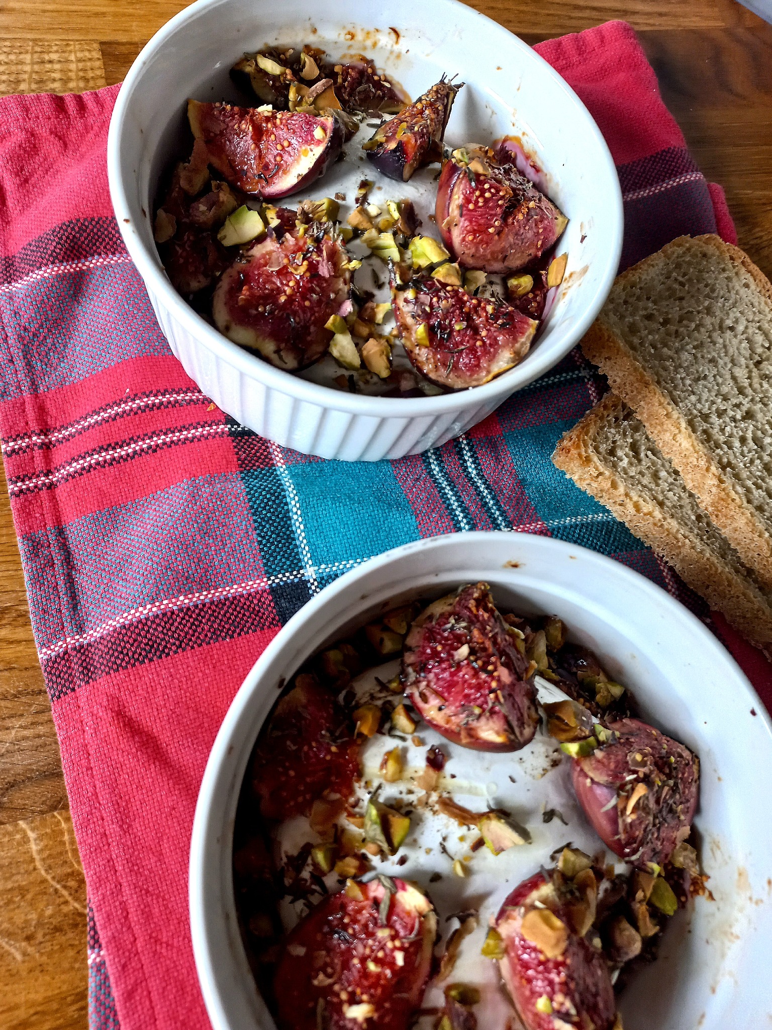 Dwa białe naczynia z pieczonymi figami posypanymi pistacjami na czerwonej, wzorzystej ściereczce obok dwóch kromek chleba na drewnianym stole.