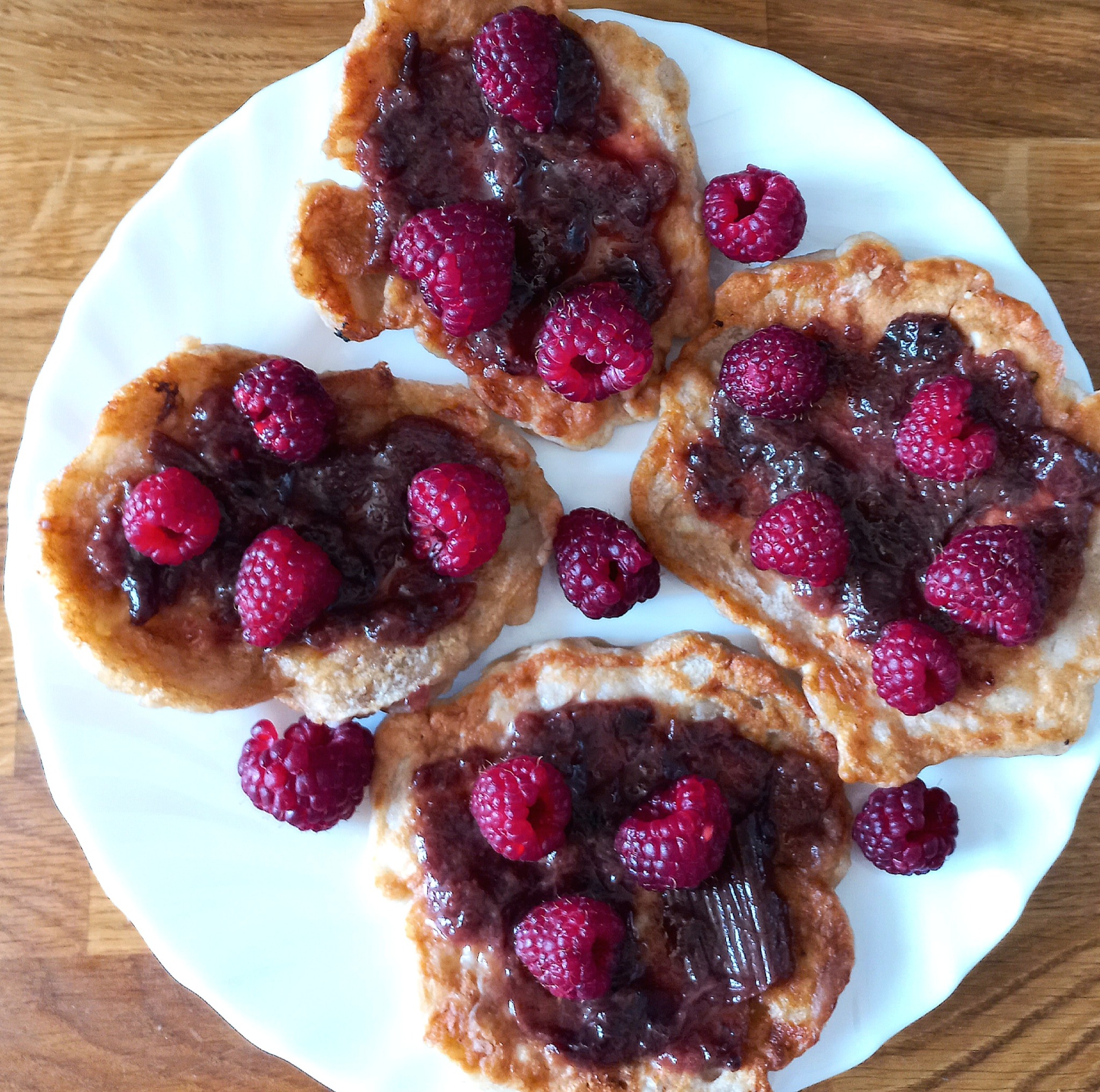 Cztery naleśniki na białym talerzu polane dżemem i udekorowane malinami, ułożone na drewnianym blacie.