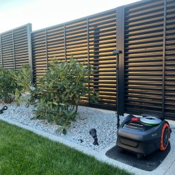 Green Home Łukasz Ogiejko - Nowoczesny ogród z robotem koszącym Navimow Efls na tle czarnego, poziomego ogrodzenia lamelowego i ozdobnych krzewów na białym żwirze, oświetlonych lampami.