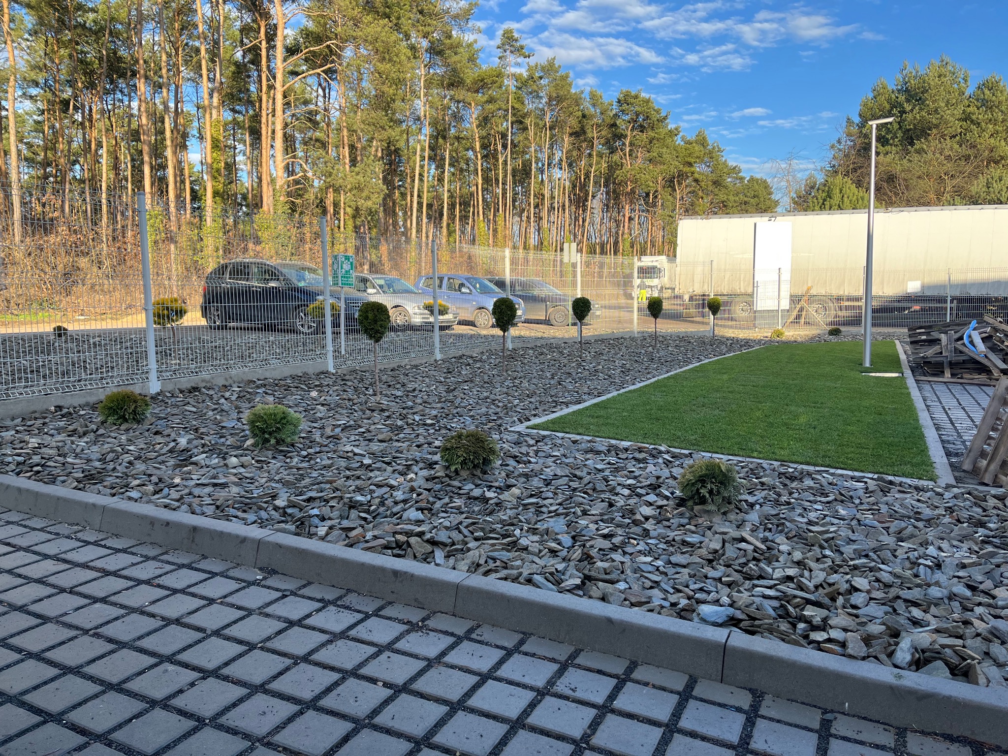 Nowoczesny ogród z kamieniami, kulistymi krzewami i fragmentem trawnika z rolki, ogrodzony siatką. W tle drzewa i budynek.