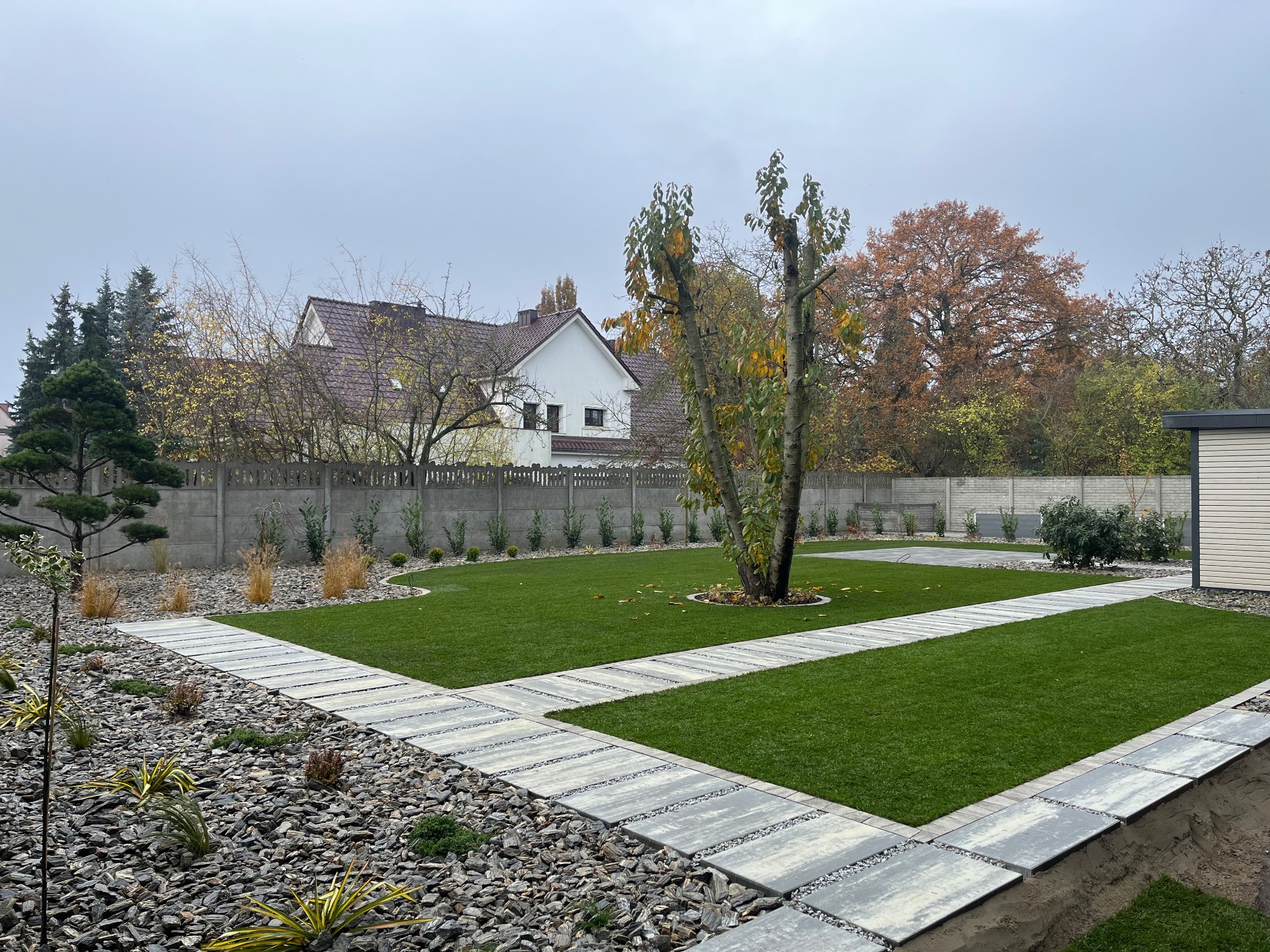 Nowoczesny ogród z równo przystrzyżonym trawnikiem, ścieżkami z płyt i ozdobnymi kamieniami. W tle widoczny dom i ogrodzenie.