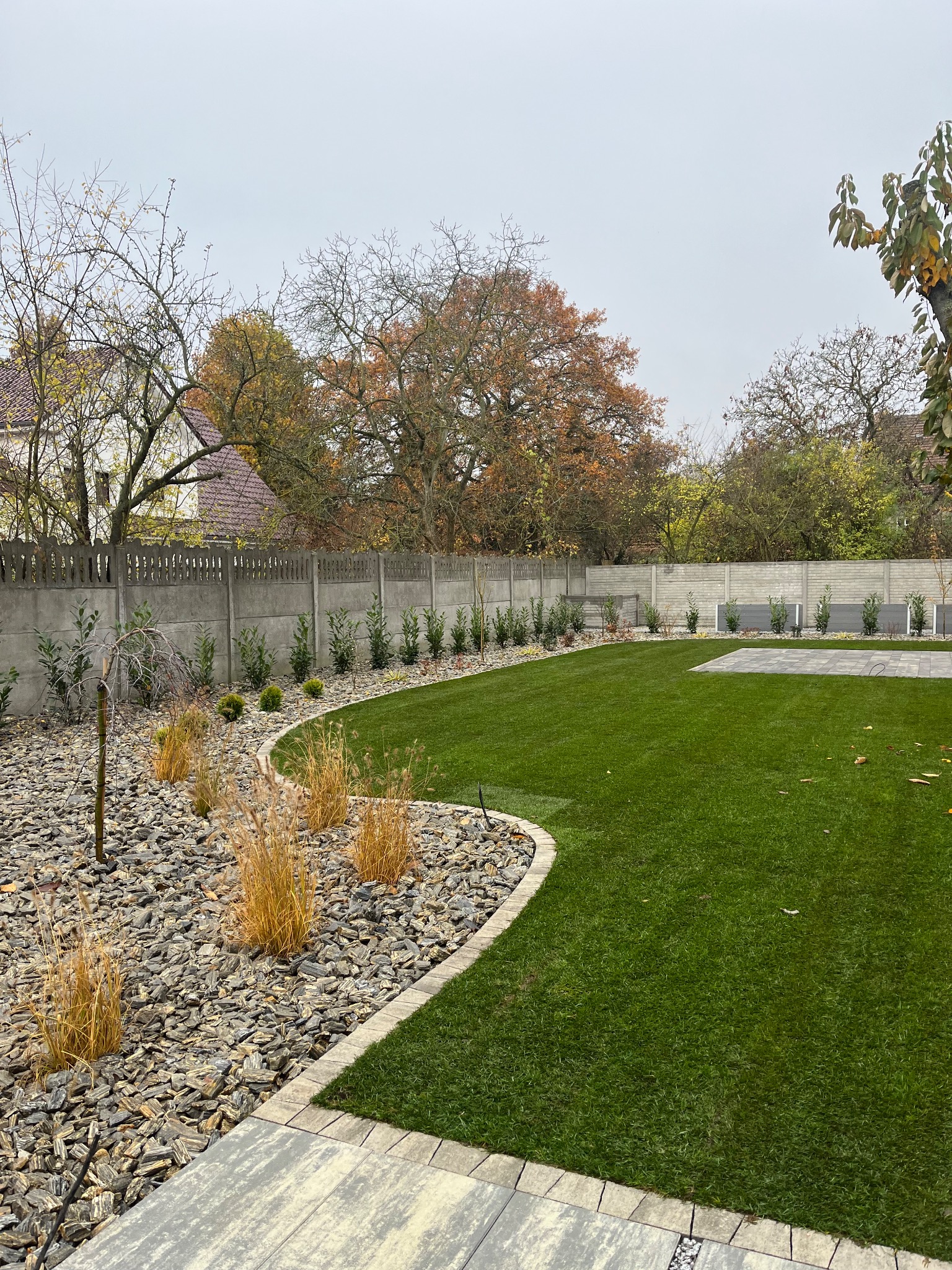 Zadbany ogród z równo przystrzyżonym trawnikiem, kamienną rabatą i ozdobnymi trawami, oddzielony betonowym płotem. Widoczne drzewa w jesiennej kolorystyce.