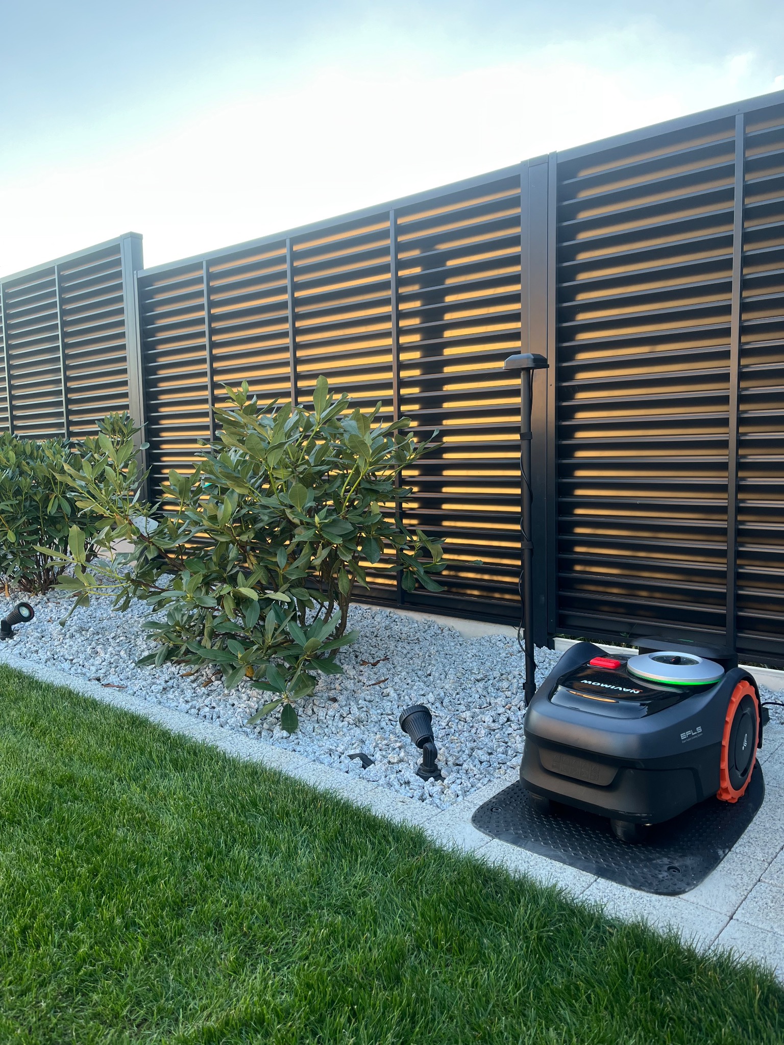 Nowoczesny ogród z robotem koszącym Navimow Efls na tle czarnego, poziomego ogrodzenia lamelowego i ozdobnych krzewów na białym żwirze, oświetlonych lampami.