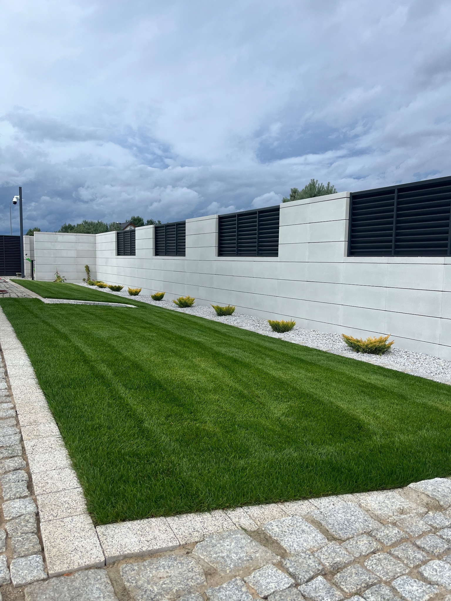 Perfekcyjnie skoszony trawnik z równymi pasami, ozdobne krzewy wzdłuż białego muru z czarnymi panelami, kamienna kostka brukowa i pochmurne niebo.