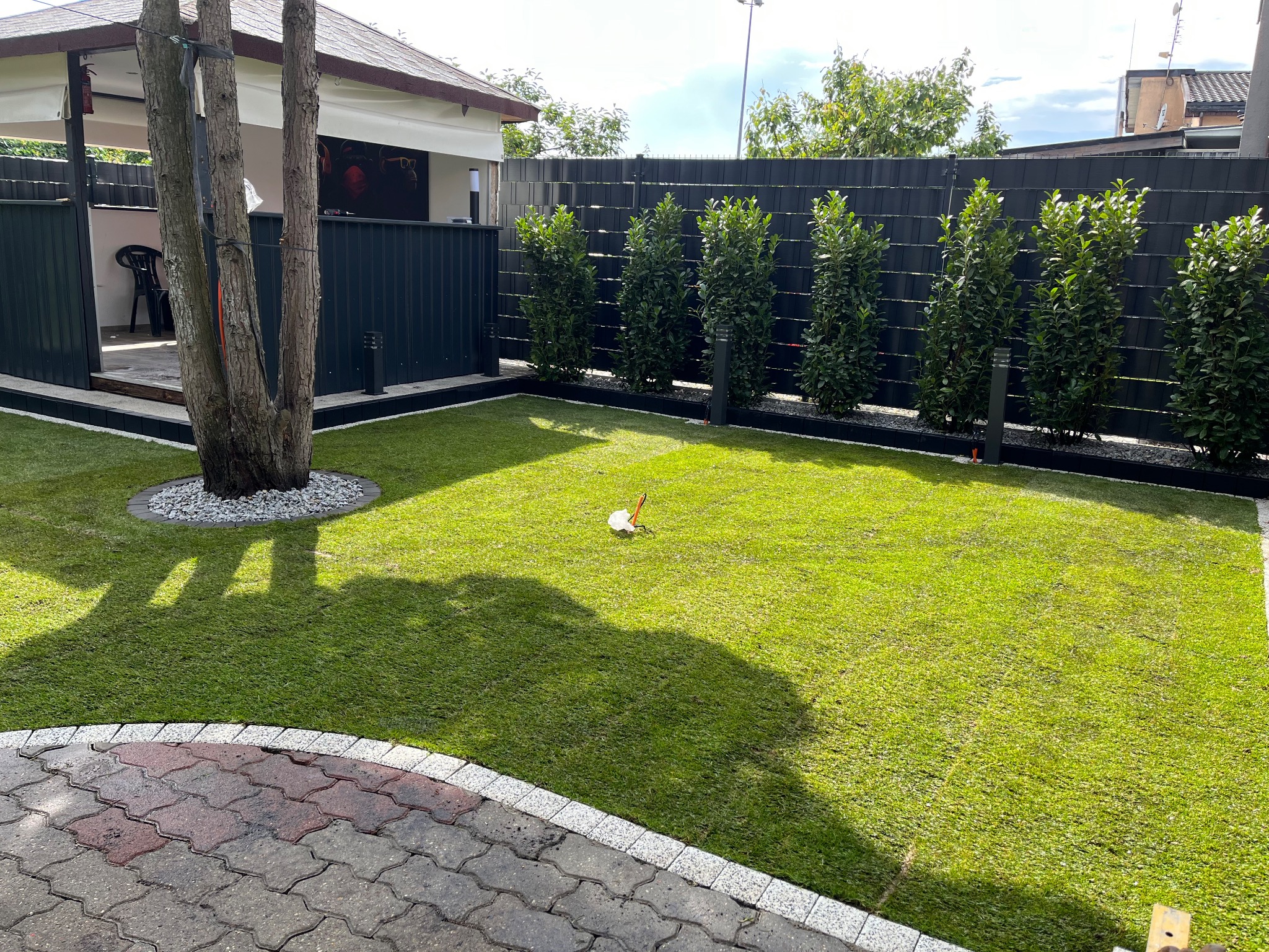 Zadbany ogród z równo przystrzyżoną trawą, obrzeżami z kamienia i rzędem krzewów wzdłuż ciemnego ogrodzenia. Widoczny fragment kostki brukowej i cień drzewa.
