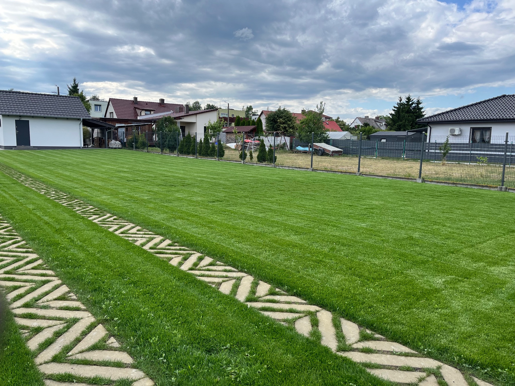 Idealnie skoszony trawnik z geometrycznym wzorem ścieżki z płyt betonowych, otoczony zielenią i zabudową jednorodzinną w tle, pod pochmurnym niebem.