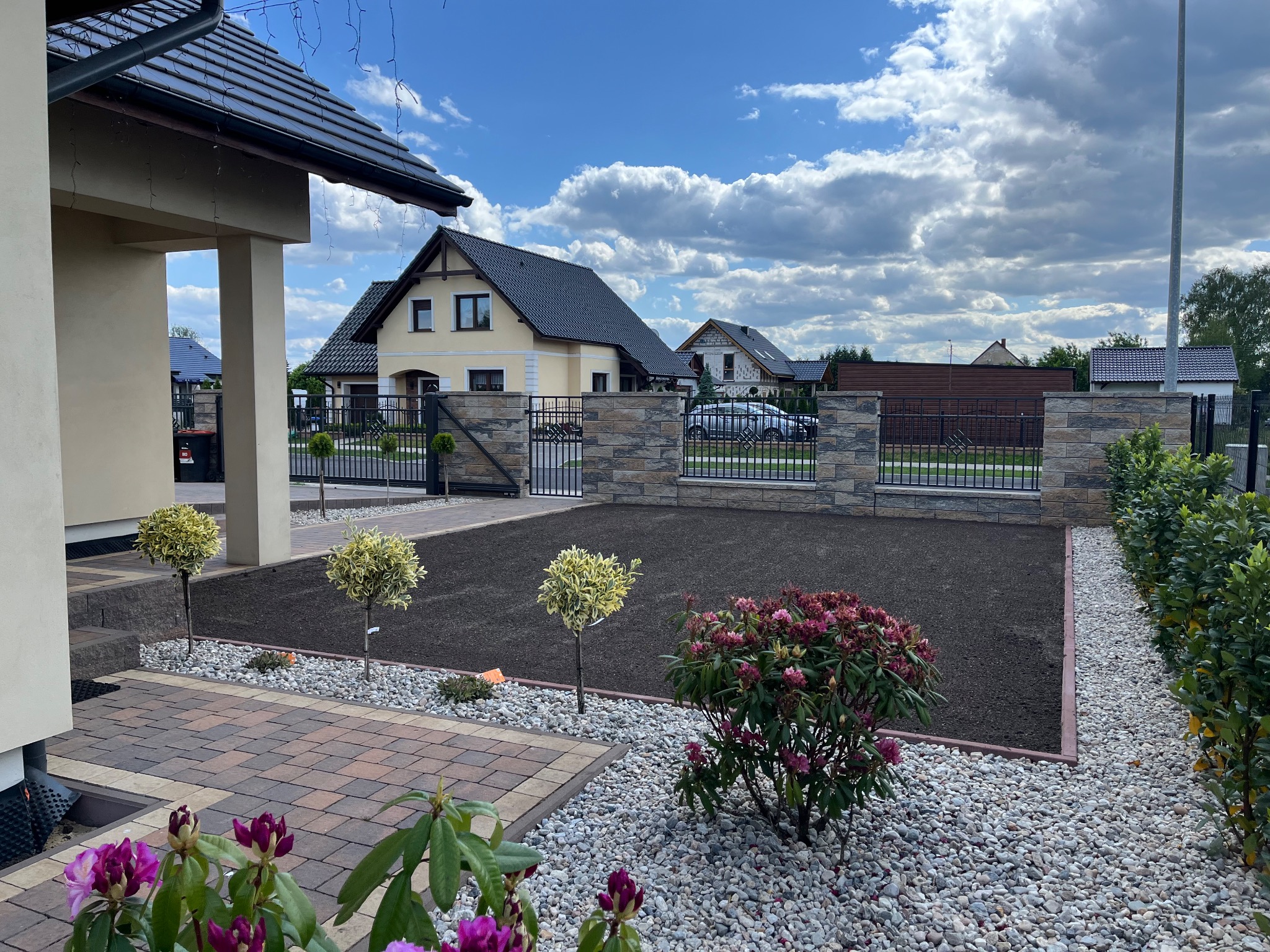 Przygotowany teren pod trawnik z rolki, obrzeża z kostki, ozdobne krzewy i drzewka na tle domu jednorodzinnego z ogrodzeniem z kamienia łupanego.