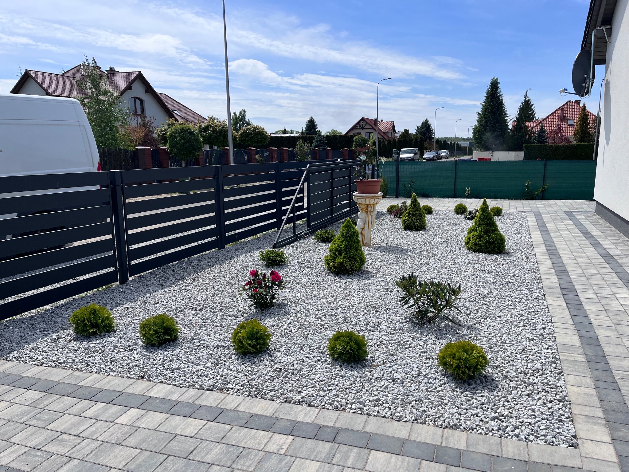 Ogród z białym żwirem, niskimi krzewami i rzeźbą z donicą. Nowoczesne ogrodzenie panelowe i kostka brukowa. Słoneczny dzień, błękitne niebo.