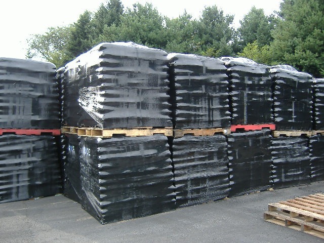 Stosy czarnych, foliowanych palet z towarem na betonowym placu, z zielenią drzew w tle.