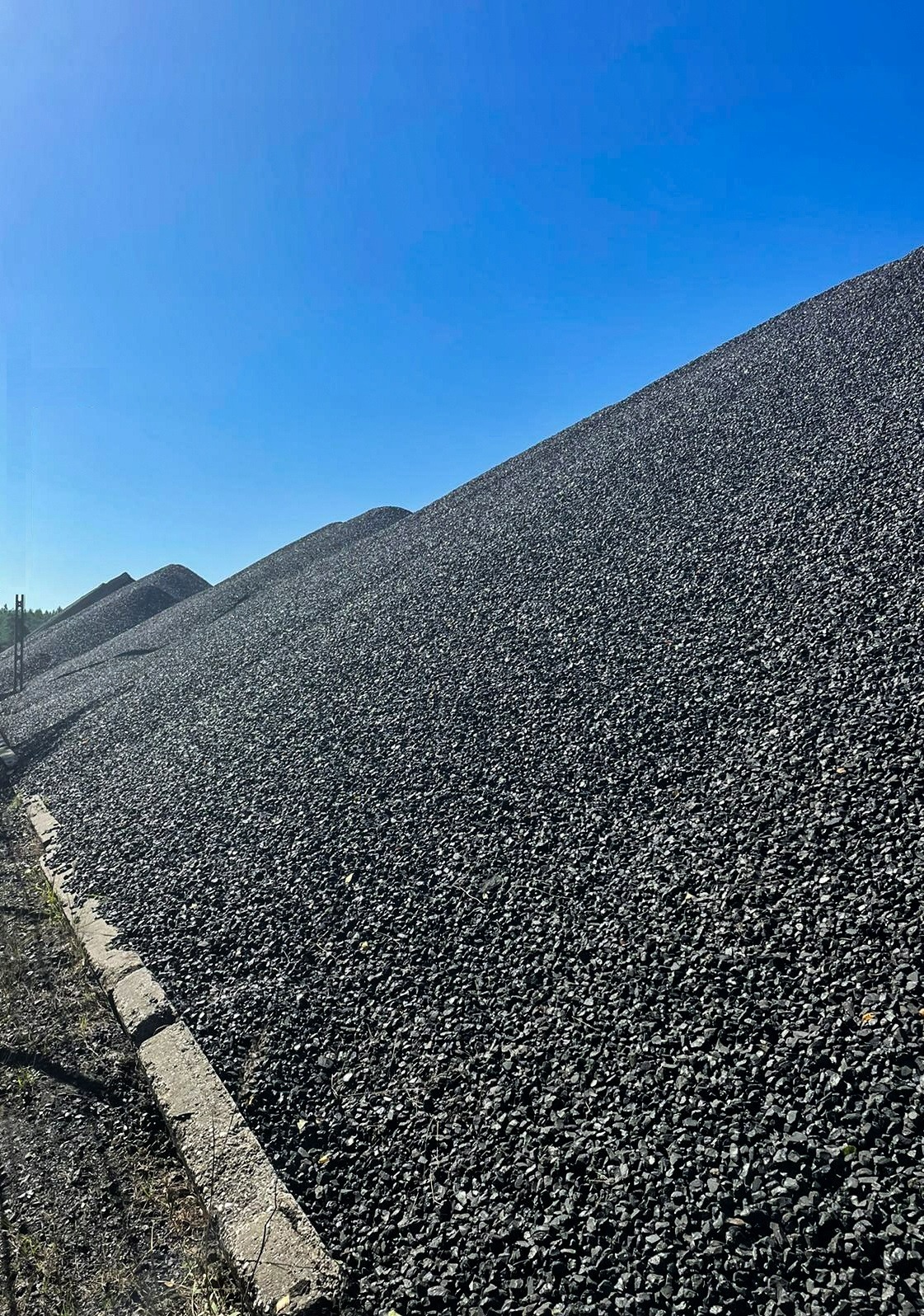 Góra czarnego węgla kamiennego pod błękitnym niebem, widoczny betonowy krawężnik u podstawy.