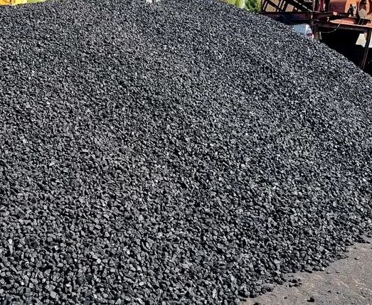 Góra czarnego ekogroszku na placu składowym, z widocznym sprzętem budowlanym w tle.