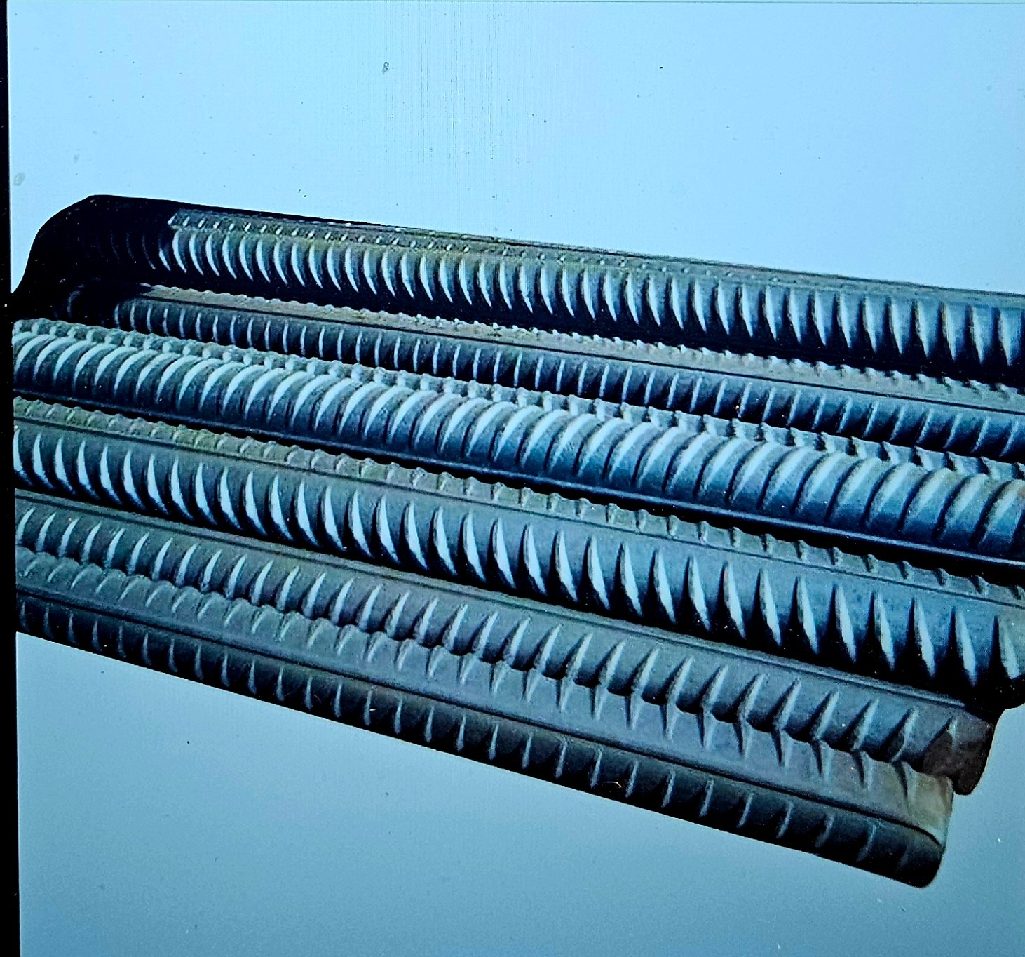 Stalowe pręty z żebrowaniem ułożone równolegle, widoczne pod kątem, na gładkiej powierzchni w chłodnym oświetleniu.