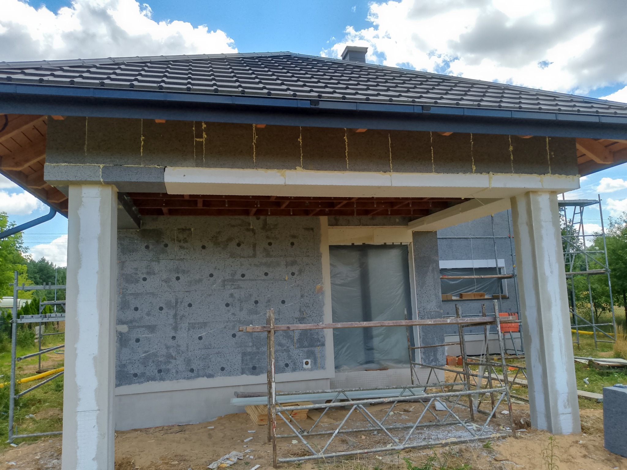 Dom w trakcie ocieplania: szara elewacja z widoczną warstwą izolacyjną, białe kolumny werandy, rusztowanie i przygotowane materiały budowlane.