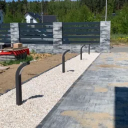 Nowoczesne ogrodzenie panelowe z szarej kostki łupanej i poziomych, metalowych przęseł, uzupełnione ścieżką z białego grysu i niskimi lampami w kształcie łuku, widoczne materiały budowlane przed...