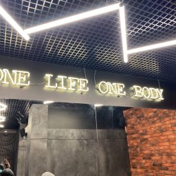 IWOPOWER Ivan Popynchuk - Neonowy napis 'ONE LIFE ONE BODY' pod sufitem z oświetleniem rastrowym w pomieszczeniu z ceglaną ścianą i szarą okładziną.