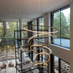 Wysoki hol w Zielonej Górze z nowoczesnym, spiralnym żyrandolem LED z metalowymi obręczami, widok z góry na rusztowanie i pracownika podczas montażu oświetlenia.