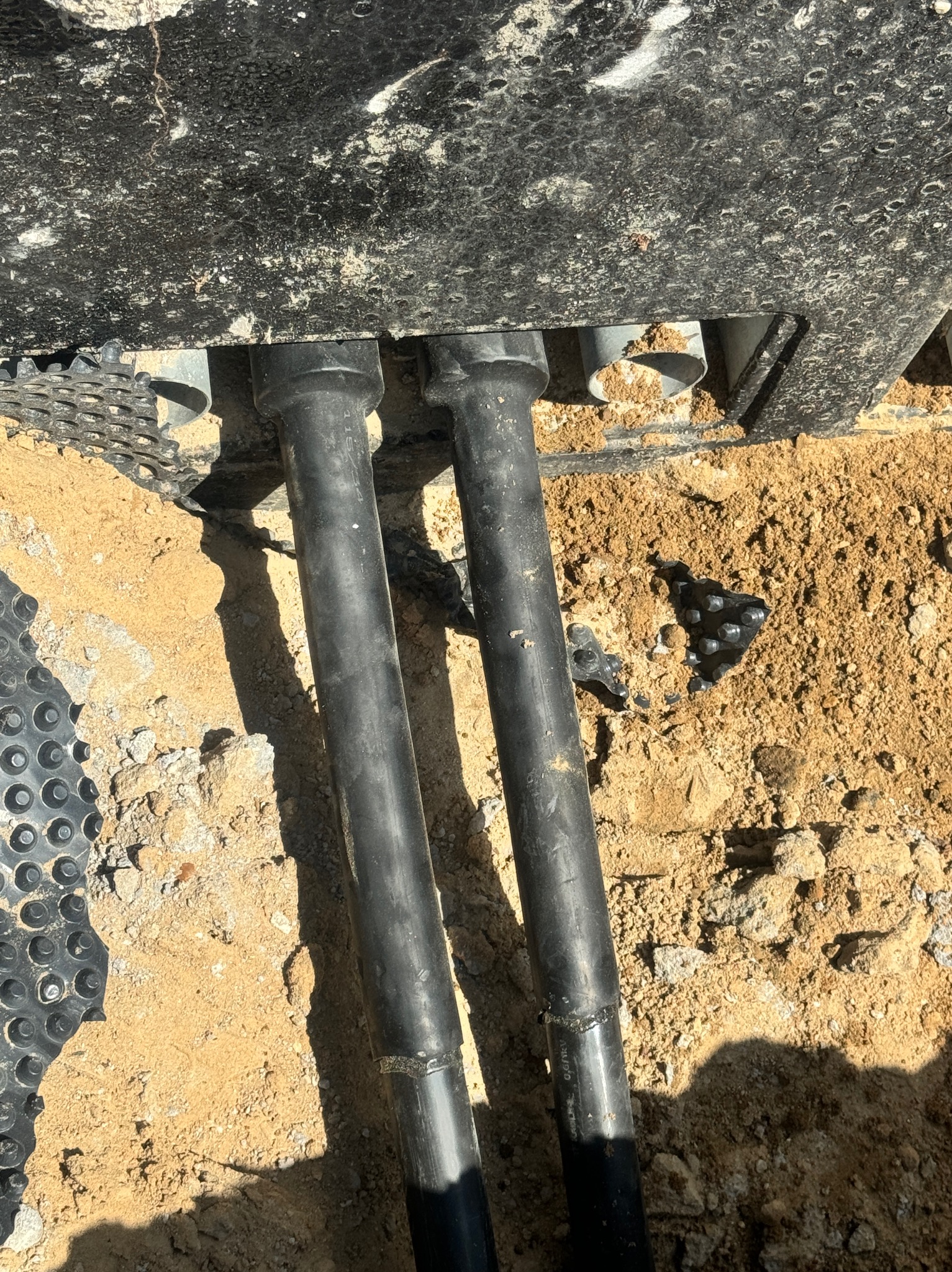 Dwa czarne, karbowane przewody w ziemi, wychodzące spod czarnej skrzynki z widocznymi szarymi rurkami, obok folia kubełkowa.