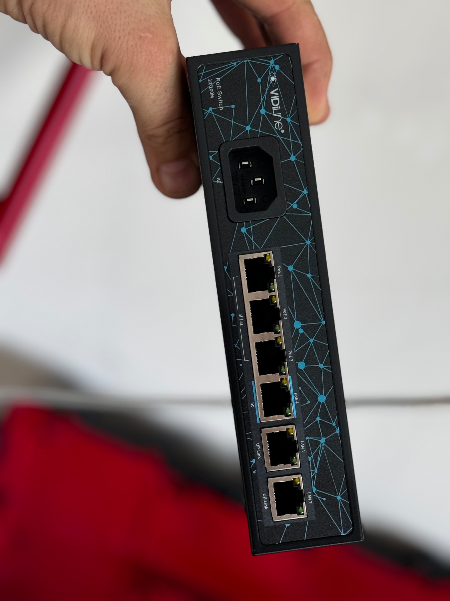 Czarny switch PoE marki Vidiline z portami Ethernet i gniazdem zasilania AC, trzymany w dłoni, widoczne porty LAN i PoE oraz wzór na obudowie.