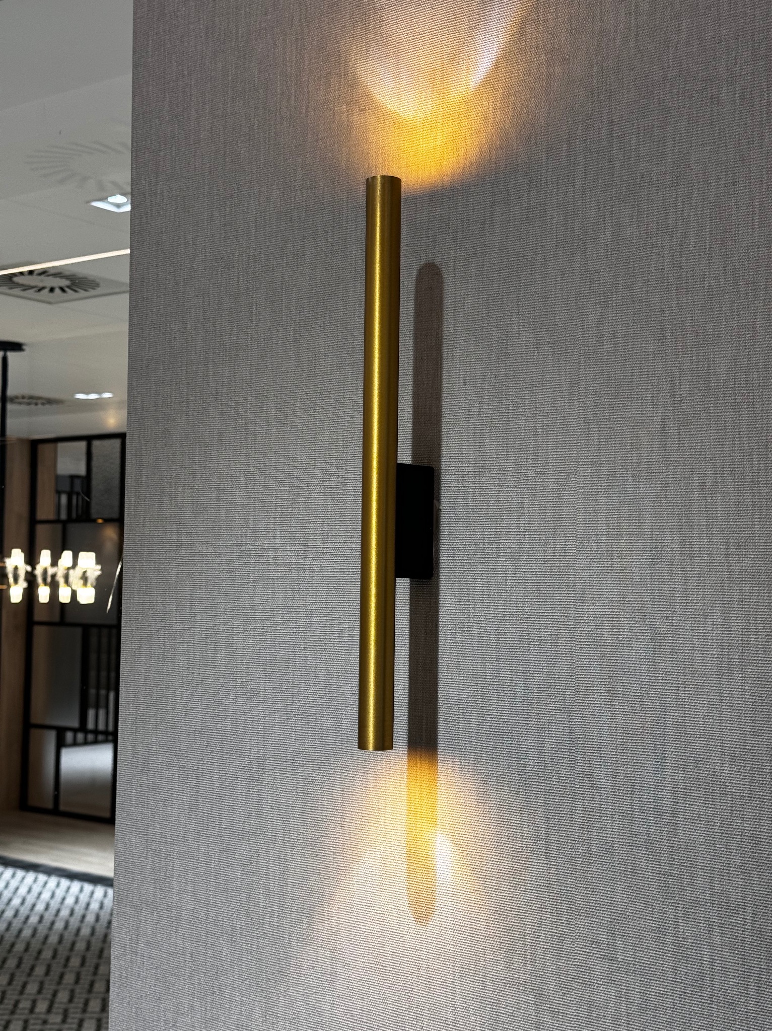 Złota, pionowa lampa ścienna o cylindrycznym kształcie, emitująca ciepłe światło w górę i w dół, zamontowana na szarej ścianie o wyraźnej fakturze, widoczny fragment pomieszczenia z wentylacją...