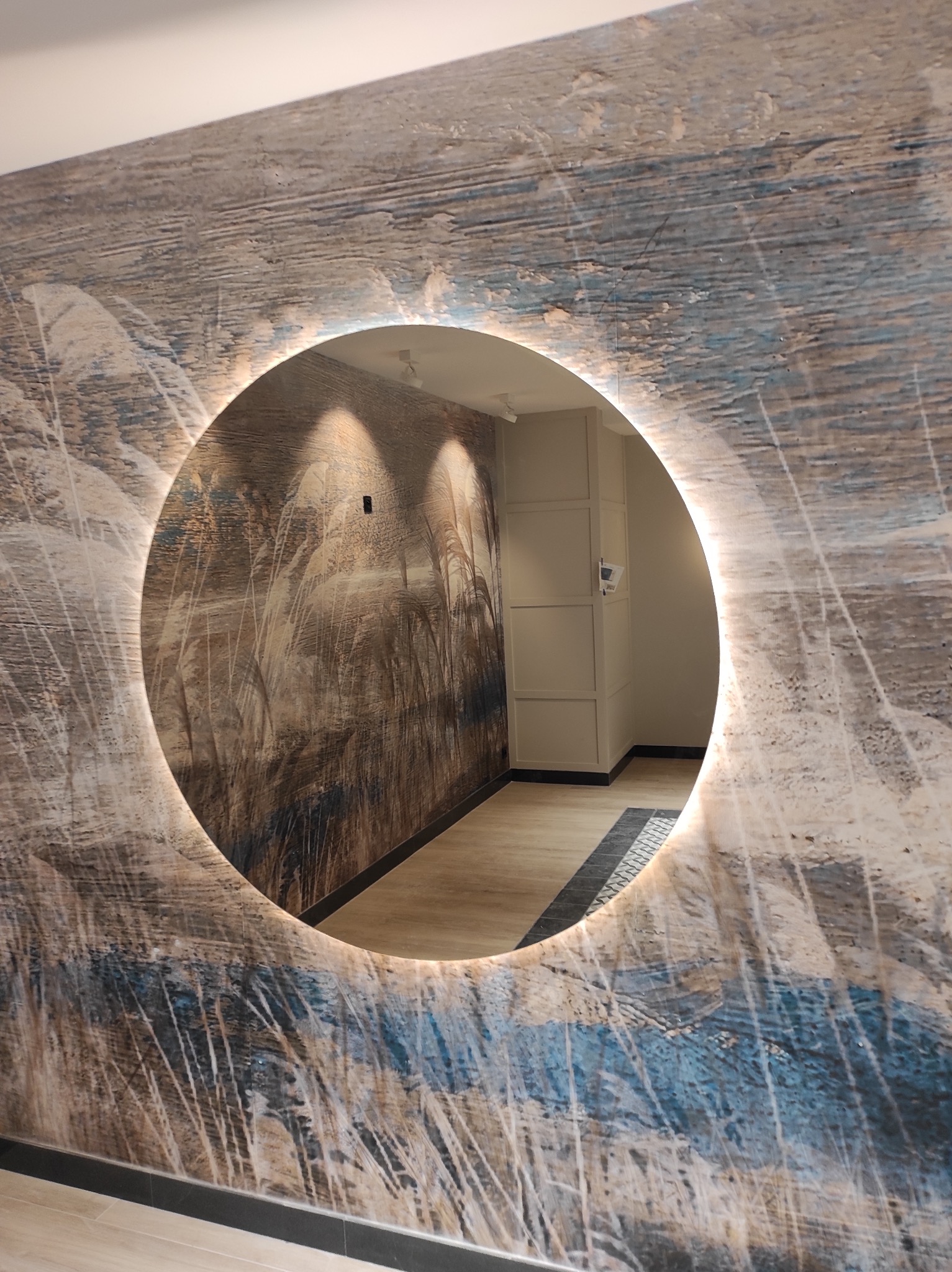 Okrągłe lustro z podświetleniem LED wkomponowane w dekoracyjną ścianę z motywem trawy, odbijające wnętrze korytarza z oświetleniem punktowym i szafą wnękową.