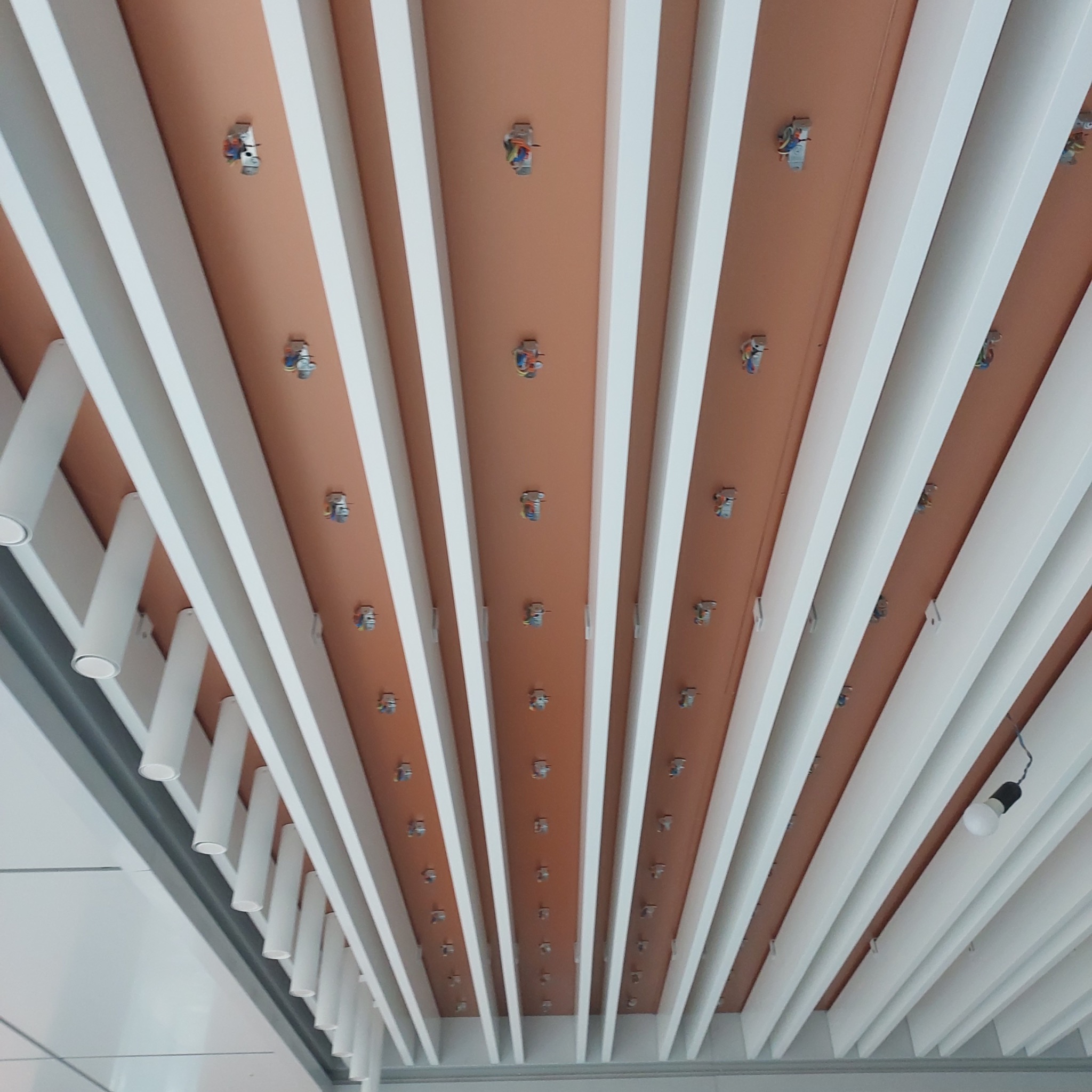 Widok z dołu na sufit z zamontowanymi uchwytami do oświetlenia, pomiędzy białymi listwami dekoracyjnymi, z widocznymi przewodami elektrycznymi.