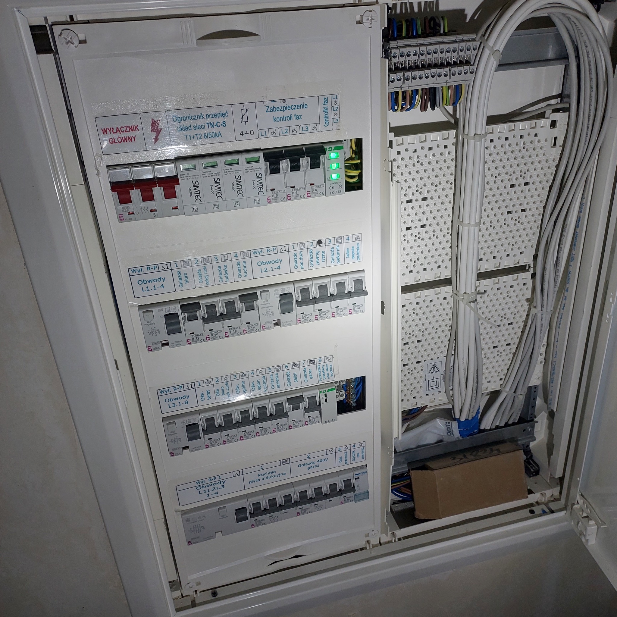 Otwarta skrzynka elektryczna z widocznymi bezpiecznikami, ogranicznikiem przepięć, zabezpieczeniem kontroli faz oraz uporządkowanymi kablami wewnątrz.
