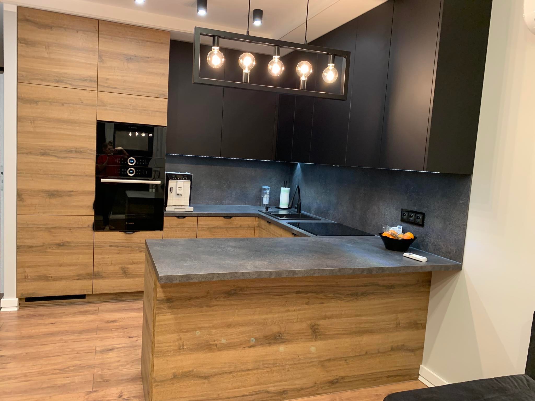 Nowoczesna kuchnia z drewnianymi szafkami i czarnymi frontami, oświetlona wiszącą lampą z żarówkami Edisona, z widoczną instalacją elektryczną gniazdek na ścianie.