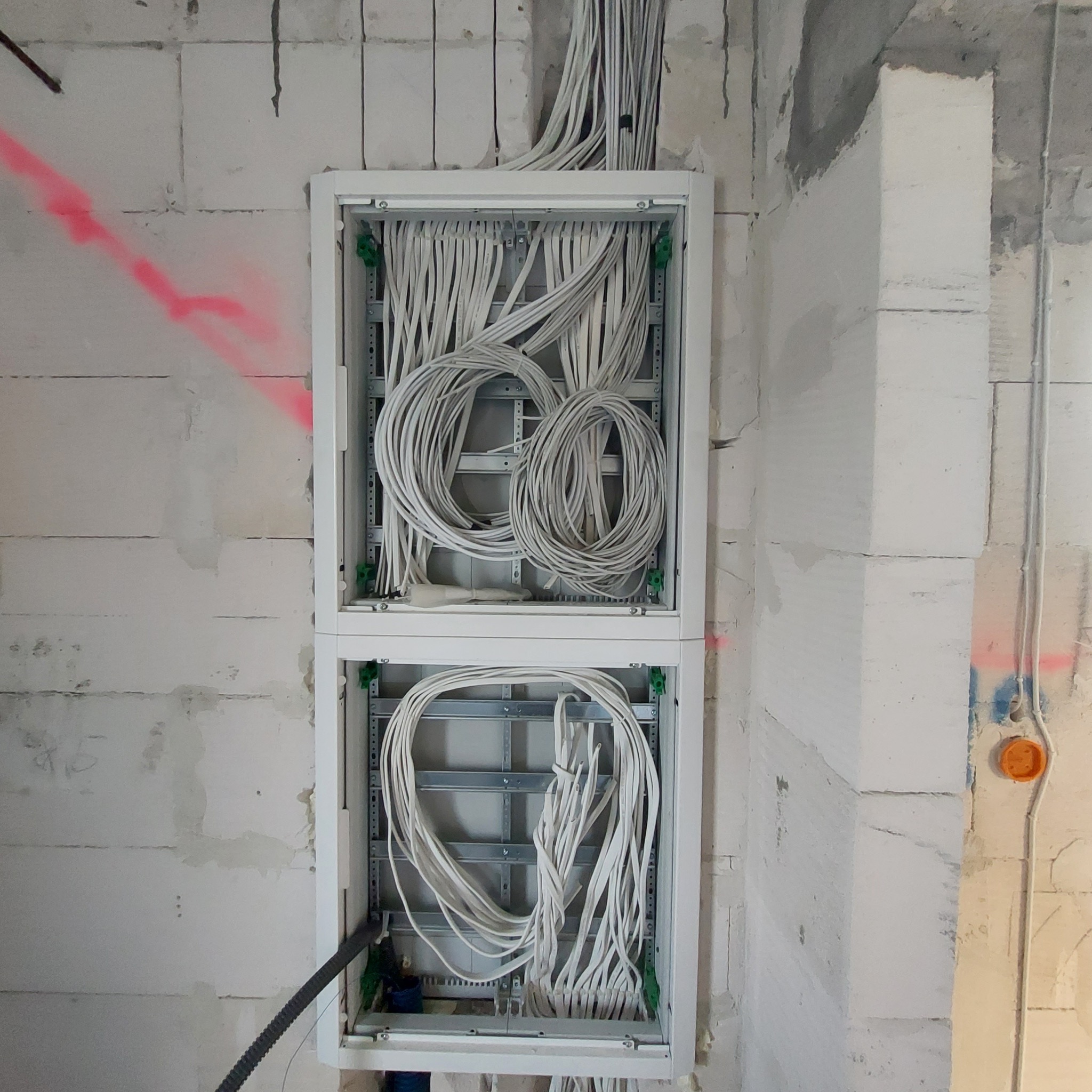 Rozdzielnica elektryczna w trakcie montażu, widoczne białe przewody ułożone w pętle wewnątrz obudowy, ściana z betonu komórkowego z widocznymi śladami po wyznaczeniu tras kabli czerwoną farbą.