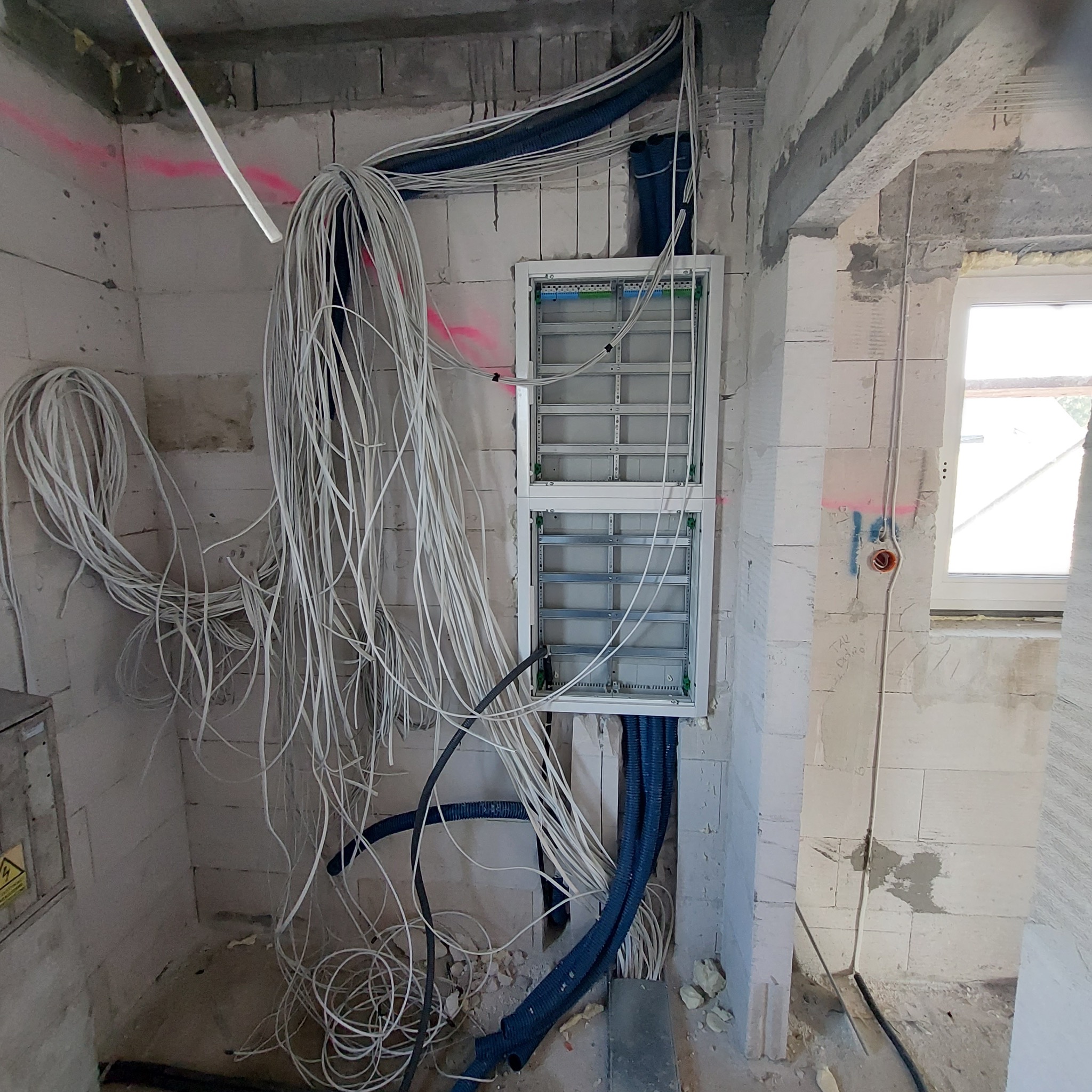 Surowy stan instalacji elektrycznej w nowym budynku: białe kable zwisają z sufitu, niebieskie rury karbowane prowadzą do otwartej skrzynki rozdzielczej na ścianie z betonu komórkowego, widoczne...
