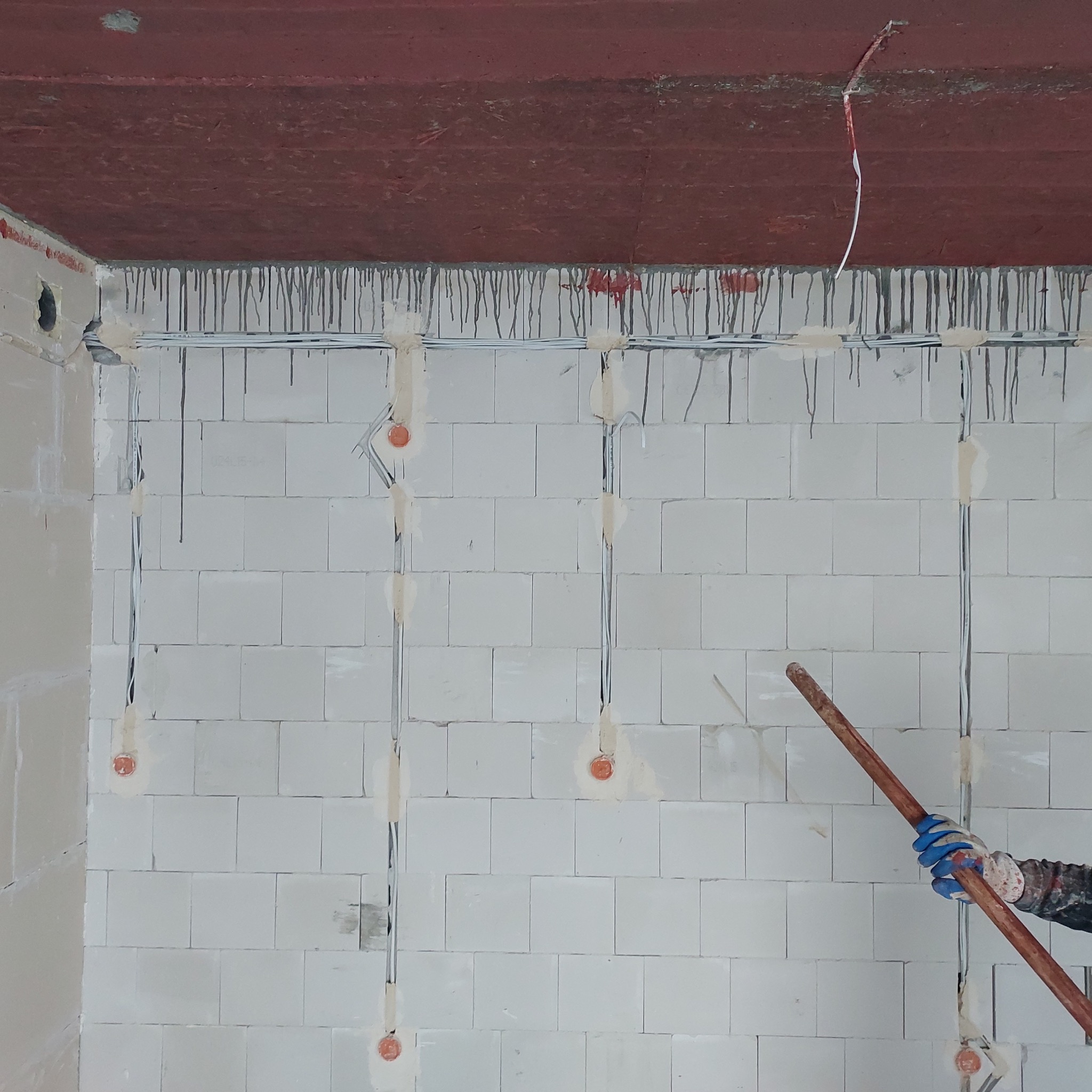 Ściana z betonu komórkowego z widocznymi przewodami elektrycznymi przytwierdzonymi taśmą, fragment dłoni w niebieskiej rękawicy trzymającej długi drewniany uchwyt