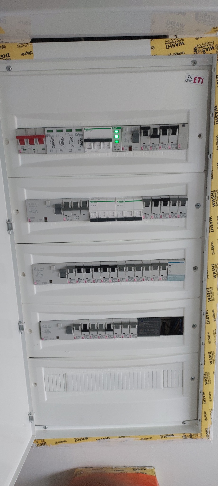 Otwarta, biała skrzynka rozdzielcza z czterema rzędami bezpieczników i wyłączników, zabezpieczona taśmą malarską na obrzeżach ściany.