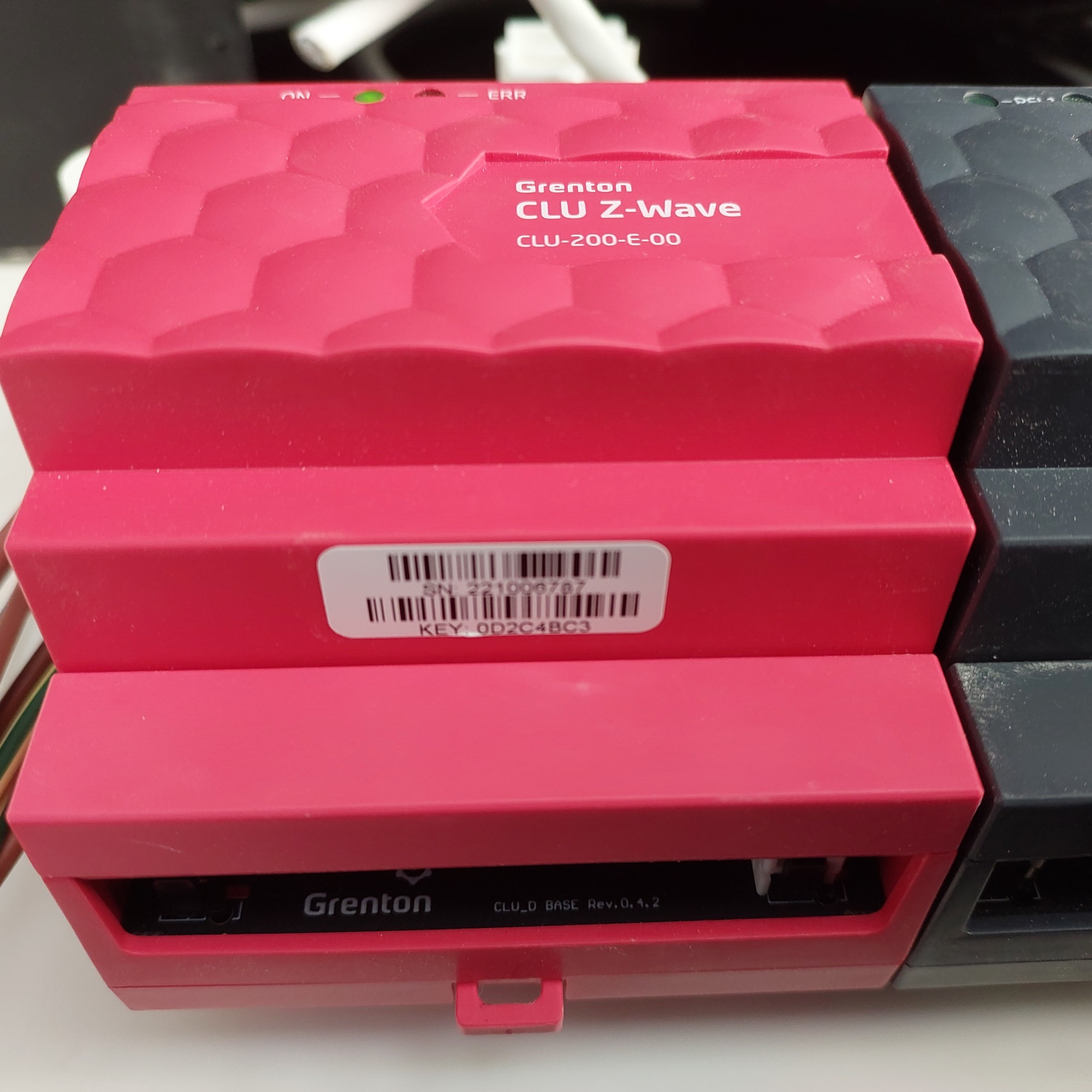 Różowy moduł sterujący Grenton CLU Z-Wave CLU-200-E-00 z widoczną etykietą z numerem seryjnym i kluczem, obok szarego modułu, na jasnym tle.